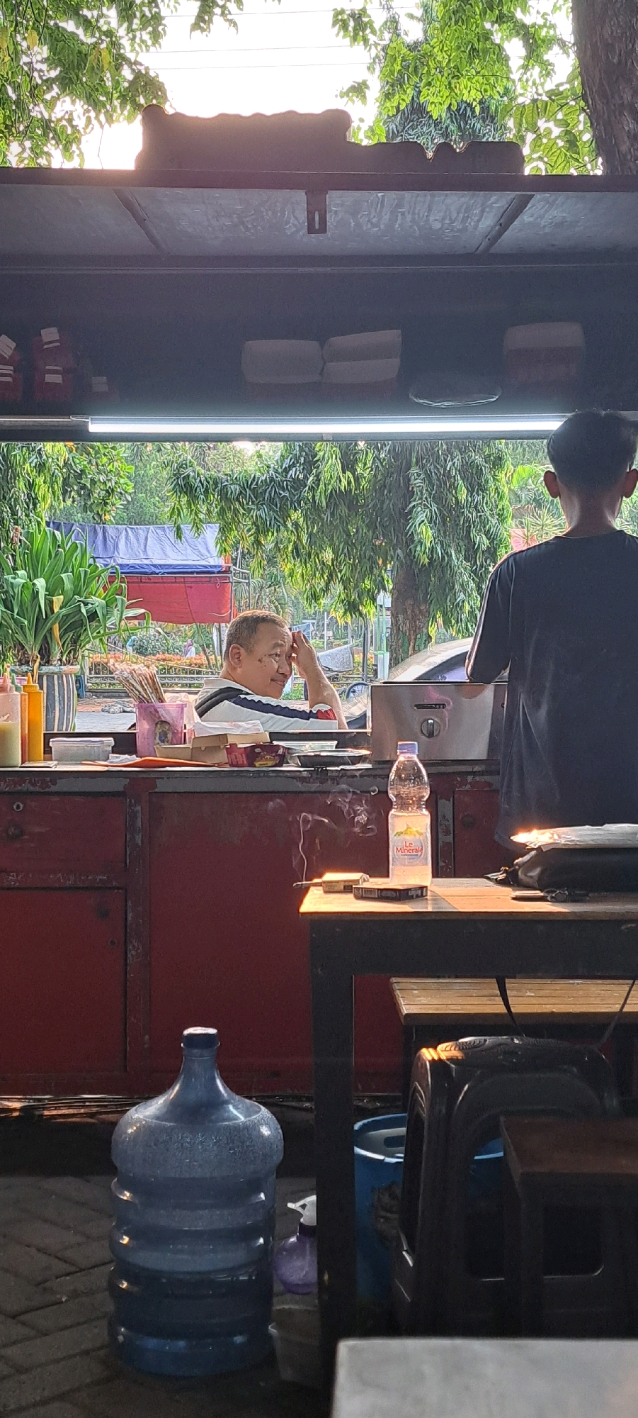 A man is sitting at a food stall, with trees and foliage visible in the background. He is wearing a striped shirt and has his hand on his forehead. Next to him is another person, wearing a dark t-shirt and facing away from the camera. On the counter, there is a bottle of water, a pack of cigarettes, and a lit cigarette emitting smoke. A large blue water jug is on the ground in front of the stall. The scene appears to be outdoors, possibly a street food vendor in Bangil, Indonesia, given the context. The lighting suggests it is late afternoon or early evening.