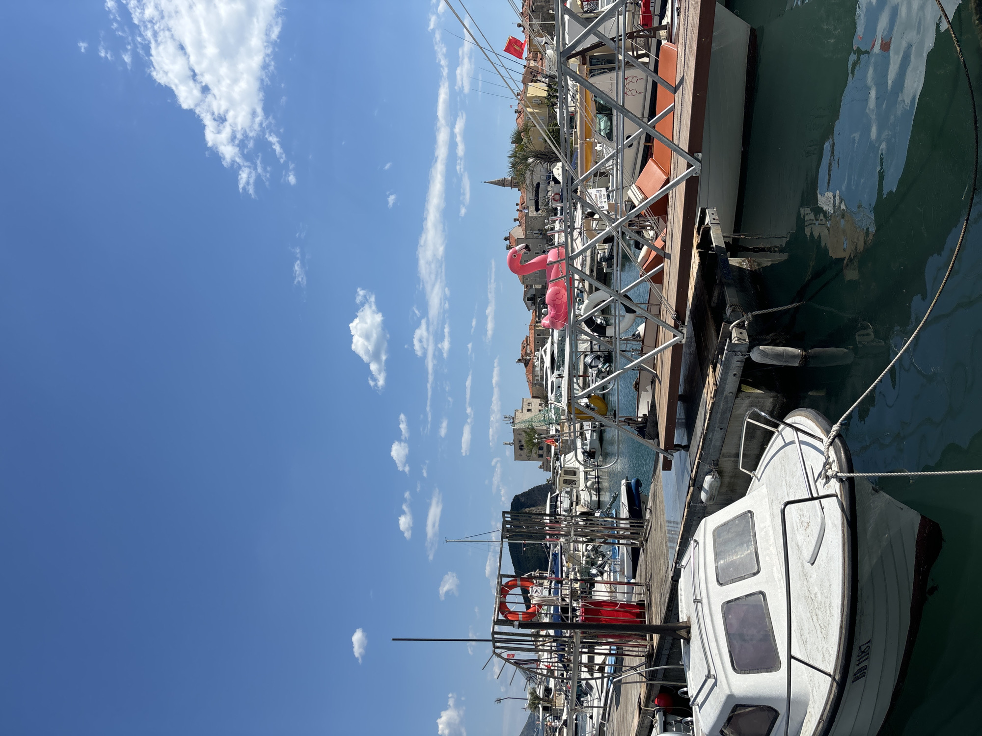 The image depicts a marina or harbor scene, likely located in Budva, Montenegro, given the presence of boats, water, and architectural elements characteristic of the region. The sky is bright blue with scattered cumulus clouds. Numerous boats of varying sizes and styles are docked along the pier, including sailboats and motorboats. A pink flamingo float is seen among the boats. In the background, there is a cluster of buildings with red-tiled roofs, which is typical of Mediterranean architecture. A church steeple is visible. The foreground features a white boat. The water is calm, reflecting the sky and surrounding boats. The image is oriented vertically.
