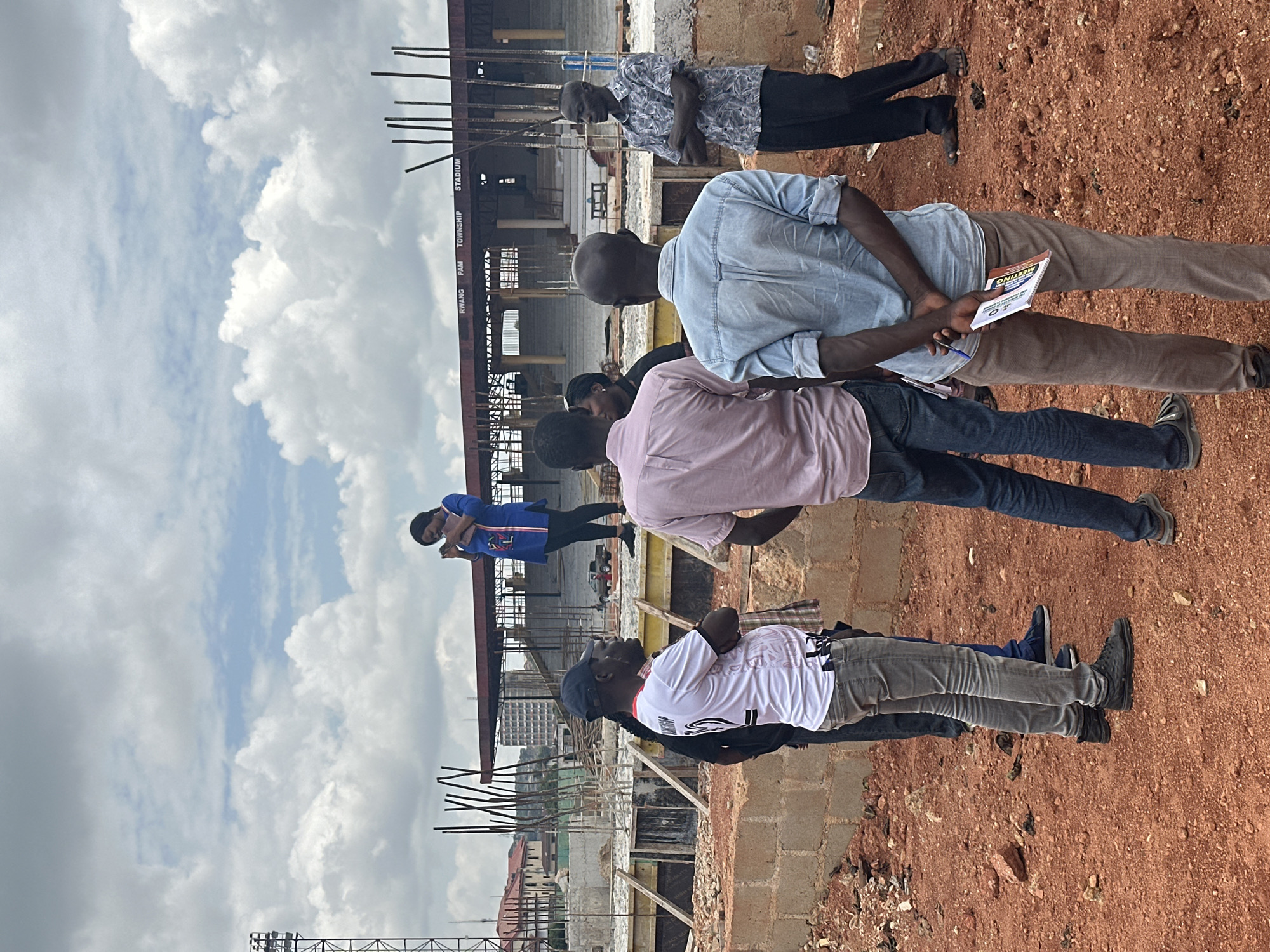 The image depicts an outdoor scene at a construction site, identified as the RWANG PAM TOWNSHIP STADIUM in Jos, Nigeria. The sky is overcast with scattered clouds. In the foreground, several men are standing on reddish-brown earth, some with their backs to the camera. One man in a white t-shirt and jeans is speaking, facing left. Another man in a light blue denim shirt and brown pants stands with his hands behind his back, holding a booklet and pen. A third man in a patterned shirt and black pants is on the right, with arms crossed. In the mid-ground, a woman in a blue and white shirt is standing on a raised platform, looking at her phone. Behind her, construction is in progress, with scaffolding, concrete structures, and rebar visible. The stadium seating area is partially constructed in the background, and some buildings and a street with a vehicle can be seen further in the distance.