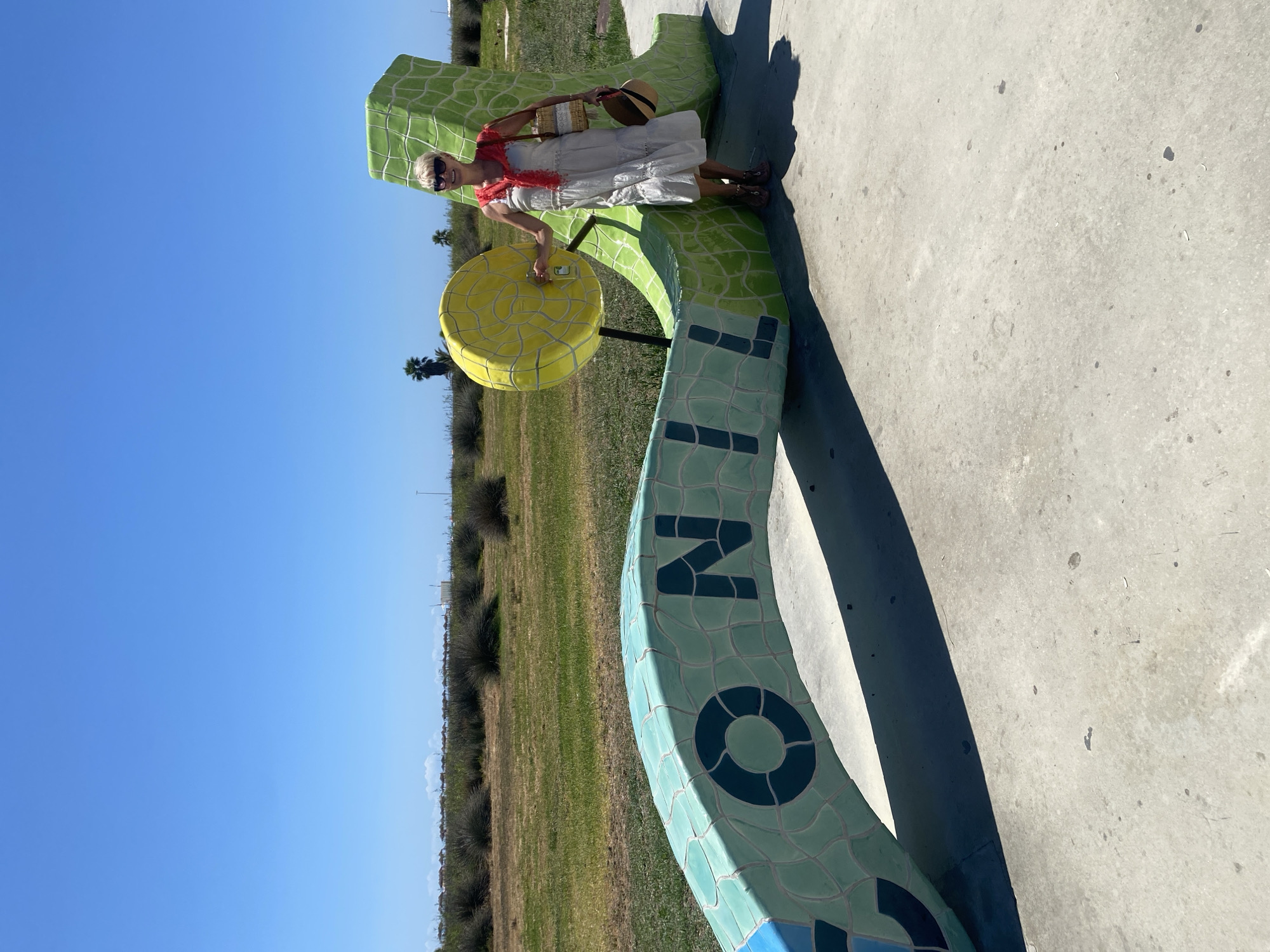 A woman stands next to a large, colorful, ceramic sculpture that spells out "Conil" in the city of Conil de la Frontera, Spain. The sculpture is green and blue, with a yellow circular element attached to it. The woman is wearing a light-colored dress, a red scarf, sunglasses, and a straw hat in her hand, along with a straw bag. She is smiling and looking at the camera. The sculpture is located outdoors on a paved surface, with grass and scrubby vegetation in the background. The sky is clear and blue. In the distance, there are rows of thatched umbrellas, suggesting a beach is nearby.