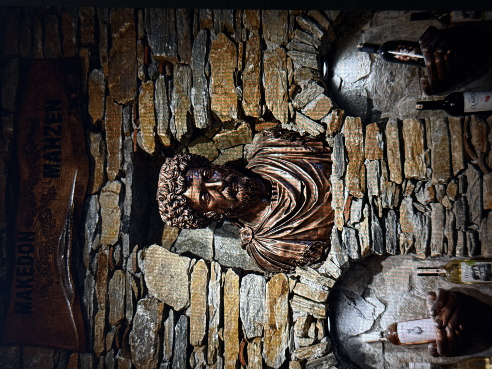 This media file depicts a dimly lit, rustic interior, possibly a cellar or a display within a historical building, given the stone wall construction and the presence of wine bottles. The primary subject is a sculpted bust of a man with curly hair and a beard, emerging from a cavity in the stone wall. The sculpture has a bronzed or copper finish, and the man wears a draped garment.
To the left of the bust, a wooden sign with carved text is visible. The text appears to read "MAKEDON MANZEN", though the lighting makes the exact inscription somewhat indistinct.
Several wine bottles are placed on ledges within the stone structure. One bottle, a dark glass bottle, is held by a hand in the upper right portion of the image. Another bottle, a lighter colored one with a label partially visible, is positioned below the first. A third bottle, also light-colored with a label, is in the lower right, held by another hand. The labels on the bottles are difficult to read clearly, but one appears to say "HERA". The overall ambiance is one of antiquity and possibly a wine tasting or cellar display. The lighting suggests an indoor setting, possibly at night or in a naturally dark environment. There are no indications of weather or specific time of day other than the artificial lighting.