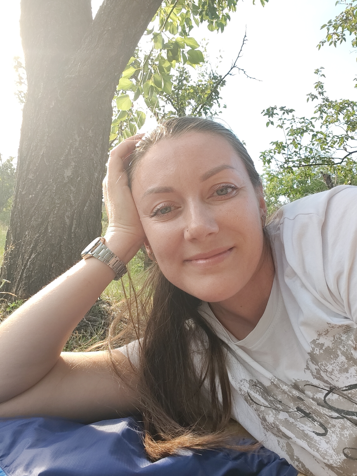 This media file is a close-up, selfie-style photograph of a woman lying down outdoors, likely in a park or natural area. She is positioned on a blue surface, possibly a mat or blanket, with her head resting on her arm. Sunlight filters through the leaves of a tree, creating a lens flare on the left side of the image, suggesting it is daytime, likely late afternoon due to the warm light.
The woman has long, brown hair and appears to be wearing a white t-shirt with a textured, abstract print. She is wearing a metallic wristwatch on her left wrist. Her right arm is bent, with her hand supporting her head. Her expression is relaxed and content, with a slight smile and her eyes looking towards the camera. The background consists of a tree trunk, green foliage, and a bright sky. There is no visible text in the image. The overall impression is one of a peaceful moment enjoying nature.