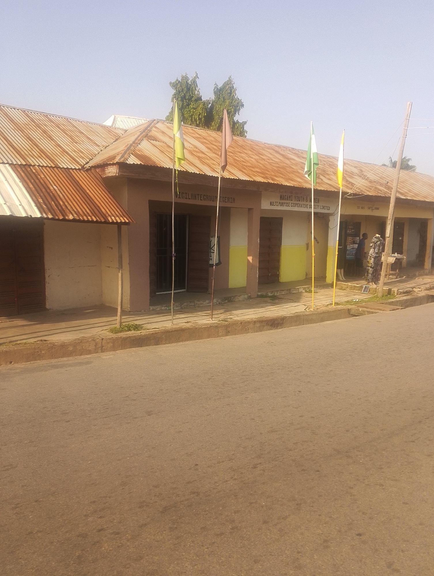A street-level, medium-shot image captures the facade of a commercial building under a clear sky. The building, with a corrugated iron roof that appears weathered and rusted, houses several businesses. The walls are painted in a pale yellow and beige hue, with some sections showing signs of wear. Three flags, two vertical and one horizontal, are prominently displayed in front of the building. The vertical flags are on poles, with the one on the left being green and white, and the one on the right being yellow. A horizontal flag, brown in color, is affixed to a pole to the left of the building.
Several storefronts are visible, with some doors closed and others open. In one open doorway, two individuals are visible. One, a person wearing a blue shirt and dark trousers, stands inside the doorway. Another individual, dressed in a patterned garment, stands outside, partially obscuring the entrance. A wooden table and some shelves are set up outside one of the shops, displaying items. The street in front of the building is paved with asphalt and shows signs of age, with some darker patches and cracks. A concrete curb separates the street from the sidewalk. The environment suggests a typical urban setting in Jalingo, Nigeria, with the building appearing to be a modest commercial establishment.