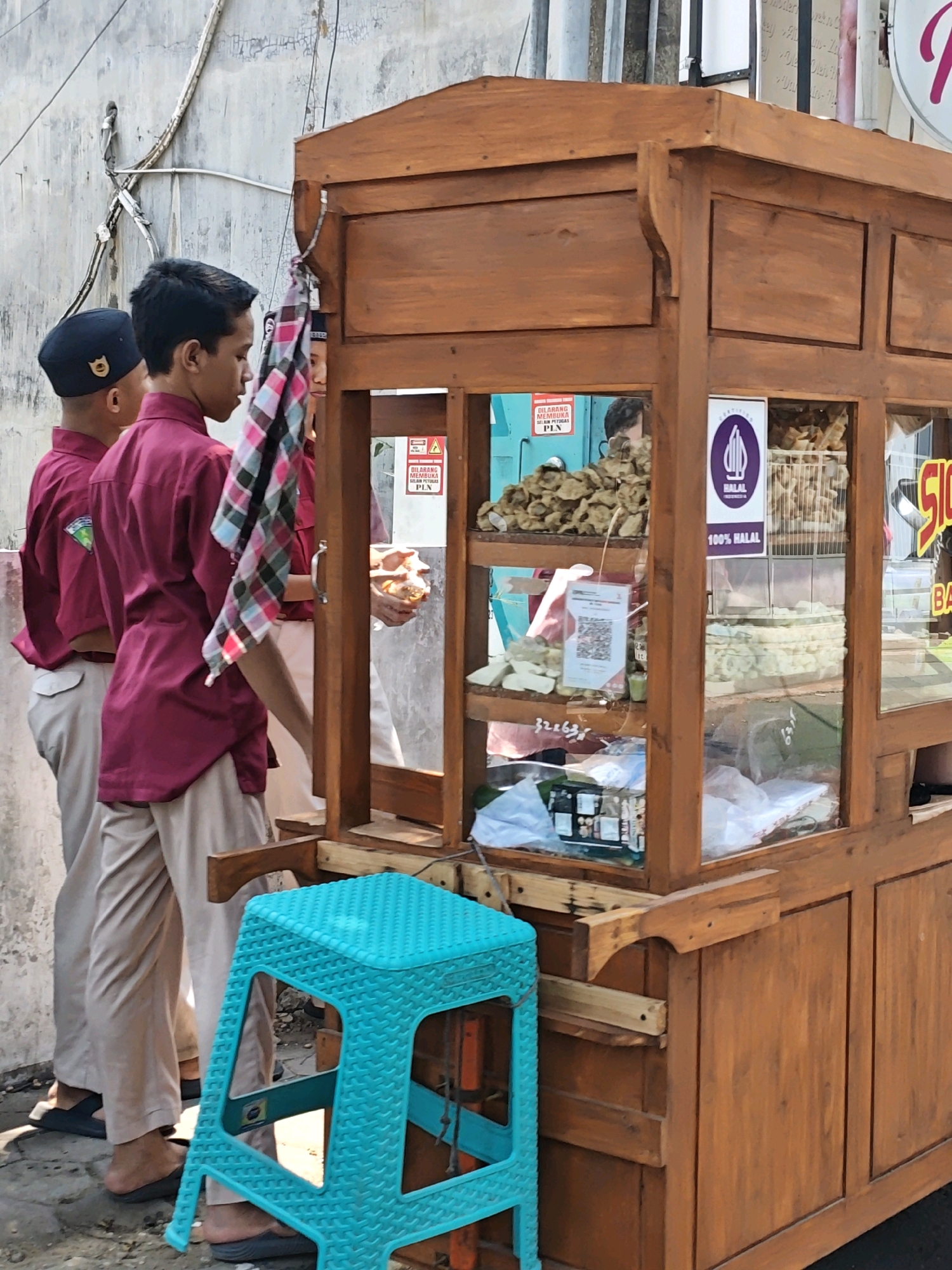 The image shows a street scene in Kediri City, Indonesia, featuring a wooden food cart and several people. Two young boys, dressed in maroon shirts and beige pants, stand near the cart. One boy wears a black peci hat. A third person, possibly another young boy or a young man, is partially visible behind the food cart, holding a piece of food. The food cart is made of dark wood and has glass display cases containing various food items, including fried snacks and what appears to be crackers. A bright turquoise plastic stool is placed in front of the cart. The background shows a textured, light-colored wall and some overhead wires. Signs on the cart indicate it is halal. The overall scene suggests a casual outdoor food vendor setting.