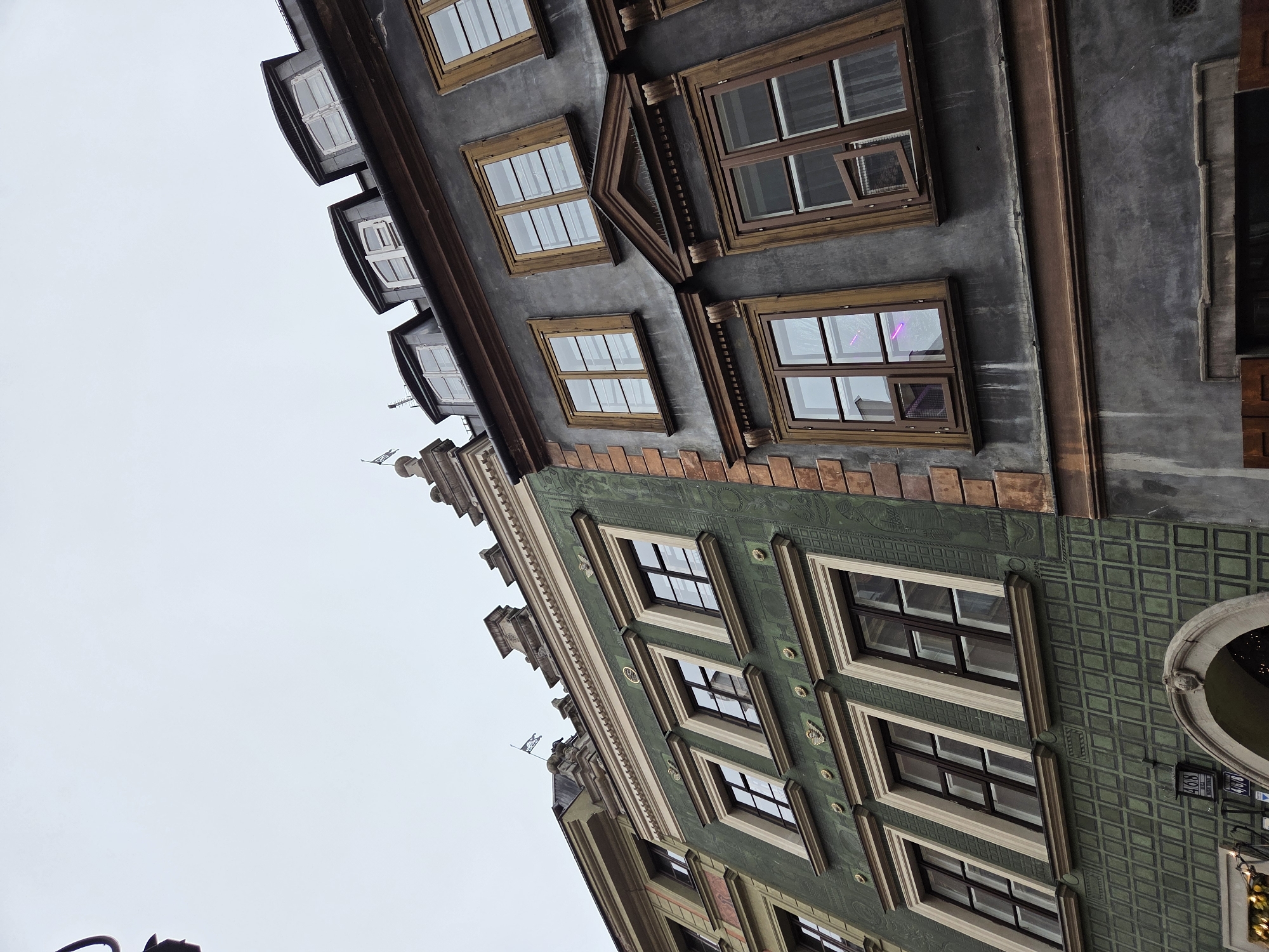 The image shows a partial view of buildings in Warsaw, Poland, taken from a low angle, with the buildings appearing to lean into the frame. The sky is overcast and gray. The buildings feature distinct architectural styles. The one on the left has a green facade with decorative tiling and rows of windows. The building on the right has a gray facade with brown window frames. There are smaller dormer windows at the top of the gray building. A street lamp post is partially visible at the bottom left corner of the image. Signage with numbers "468" and "34" is also visible on the right side of the green building, near the bottom.