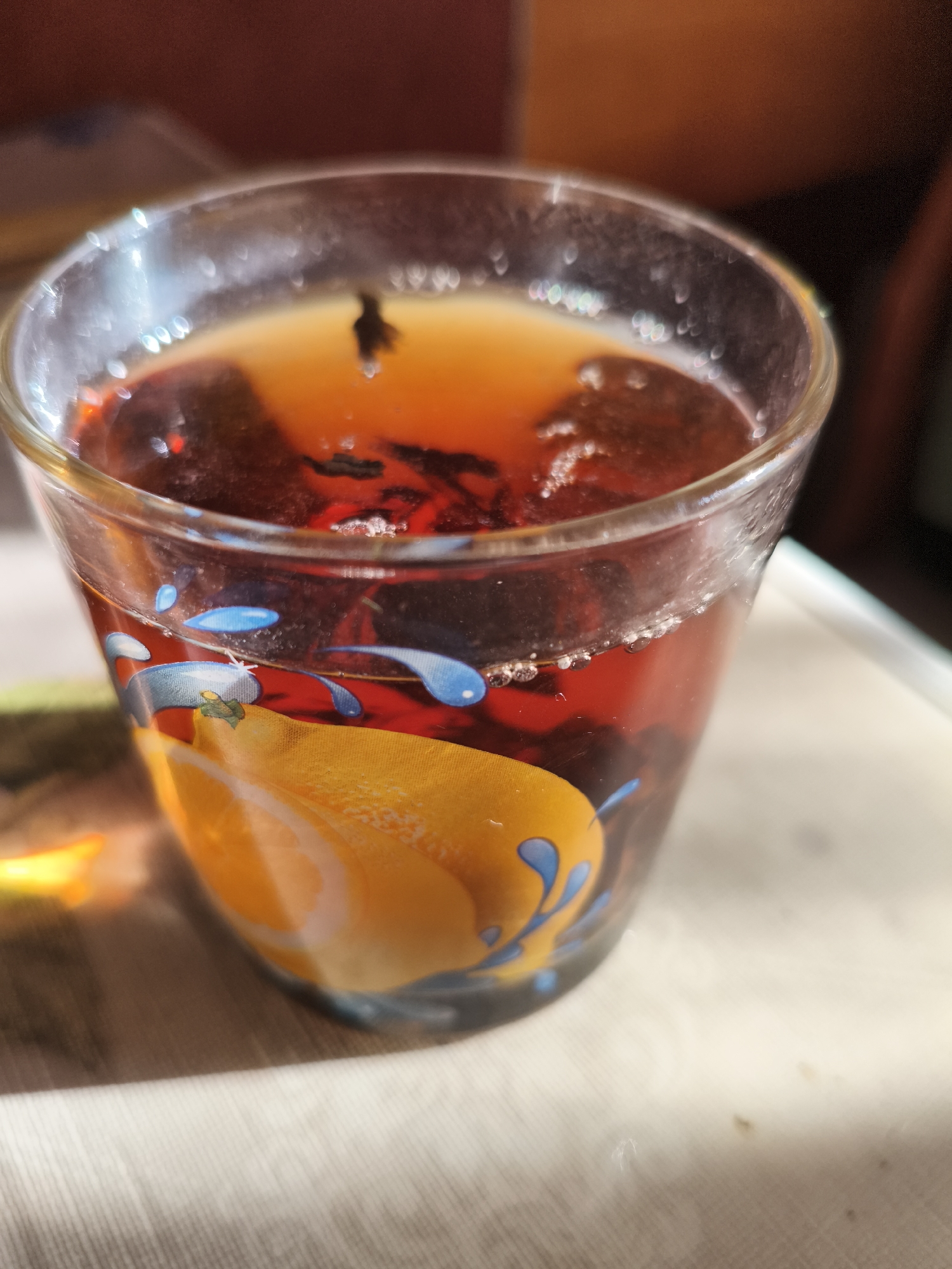 A close-up, eye-level shot shows a glass of dark red liquid, possibly tea or juice, with loose tea leaves floating on top. The glass has a decorative pattern featuring an orange slice with blue water droplets. The lighting suggests it is daytime, with sunlight casting shadows. The background is blurred, showing warm tones of wood and a lighter surface. The setting appears to be indoors, possibly a domestic or cafe environment, in the city of Ilmino, Russia. There are no people or specific actions visible.