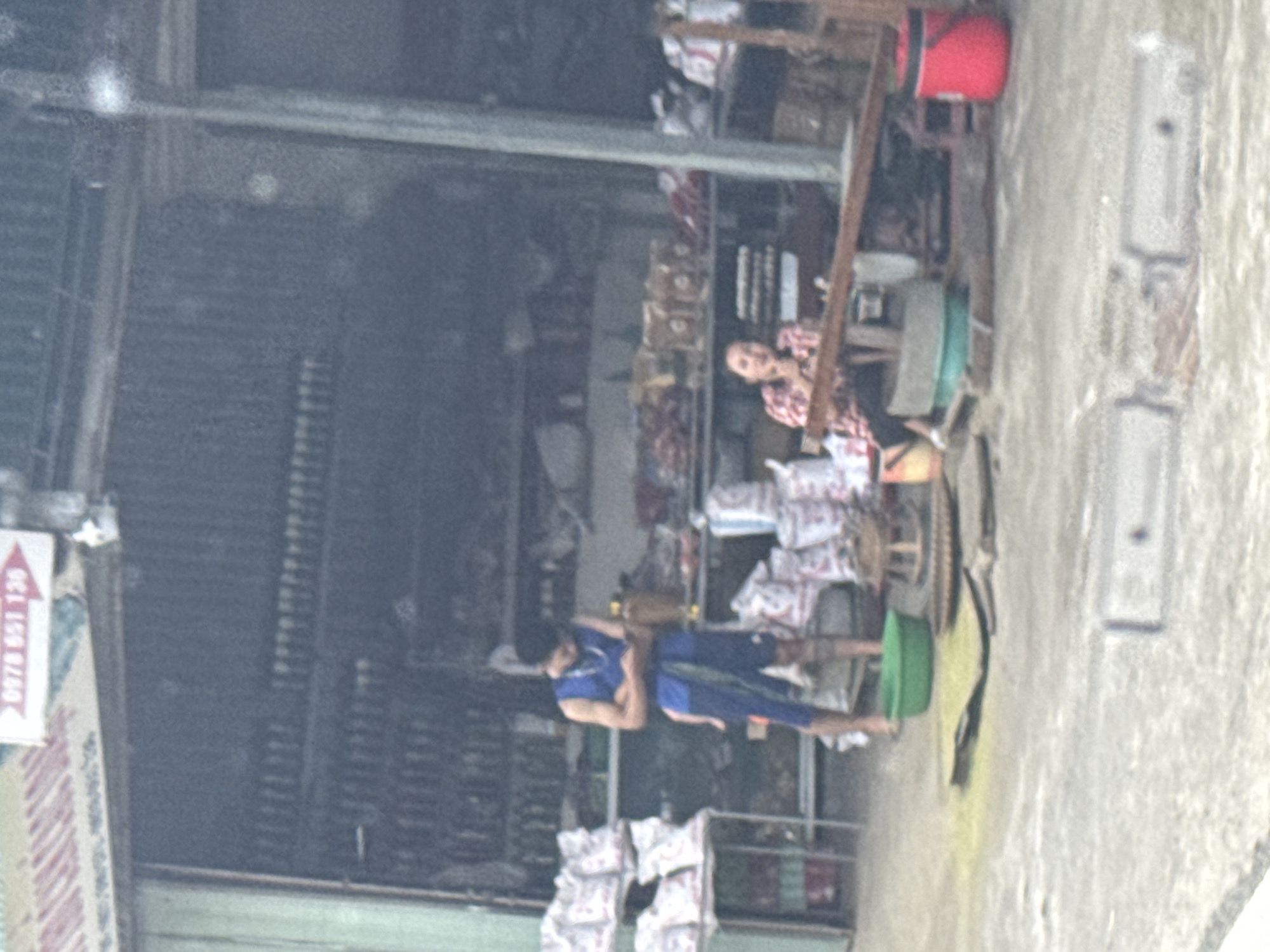 This image depicts an outdoor commercial scene in Tú Lệ, Vietnam, featuring an open-front shop or stall. The building's interior background consists of corrugated metal walls, with multiple shelves holding rows of dark-colored bottles. In the upper left portion of the image, stacks of white sacks are visible on a metal shelving unit. Above the entrance, a partial white sign with red lettering reads "0978 651 130" next to a red arrow pointing right.
Two individuals are present on a concrete ground surface outside the building. On the left, a male subject stands barefoot, wearing a blue sleeveless shirt and blue shorts, with a dark blue sash or cloth tied around his waist. He is holding a small rectangular object, likely a mobile phone, and is looking down at it. To the right, a female subject is seated directly on the ground, wearing a red and white plaid shirt, and is looking towards the left side of the frame.
Various objects are positioned on the ground between and around the individuals. These include a bright green plastic basin in the lower-left, a flat woven mat, a smaller round woven stool or tray, a larger green cylindrical container, and a prominent dark wooden beam or plank lying horizontally across the mid-ground. A red insulated bucket or container is visible in the lower right corner. Additional smaller bags and items are scattered across the ground. The overall scene suggests a retail or processing operation.