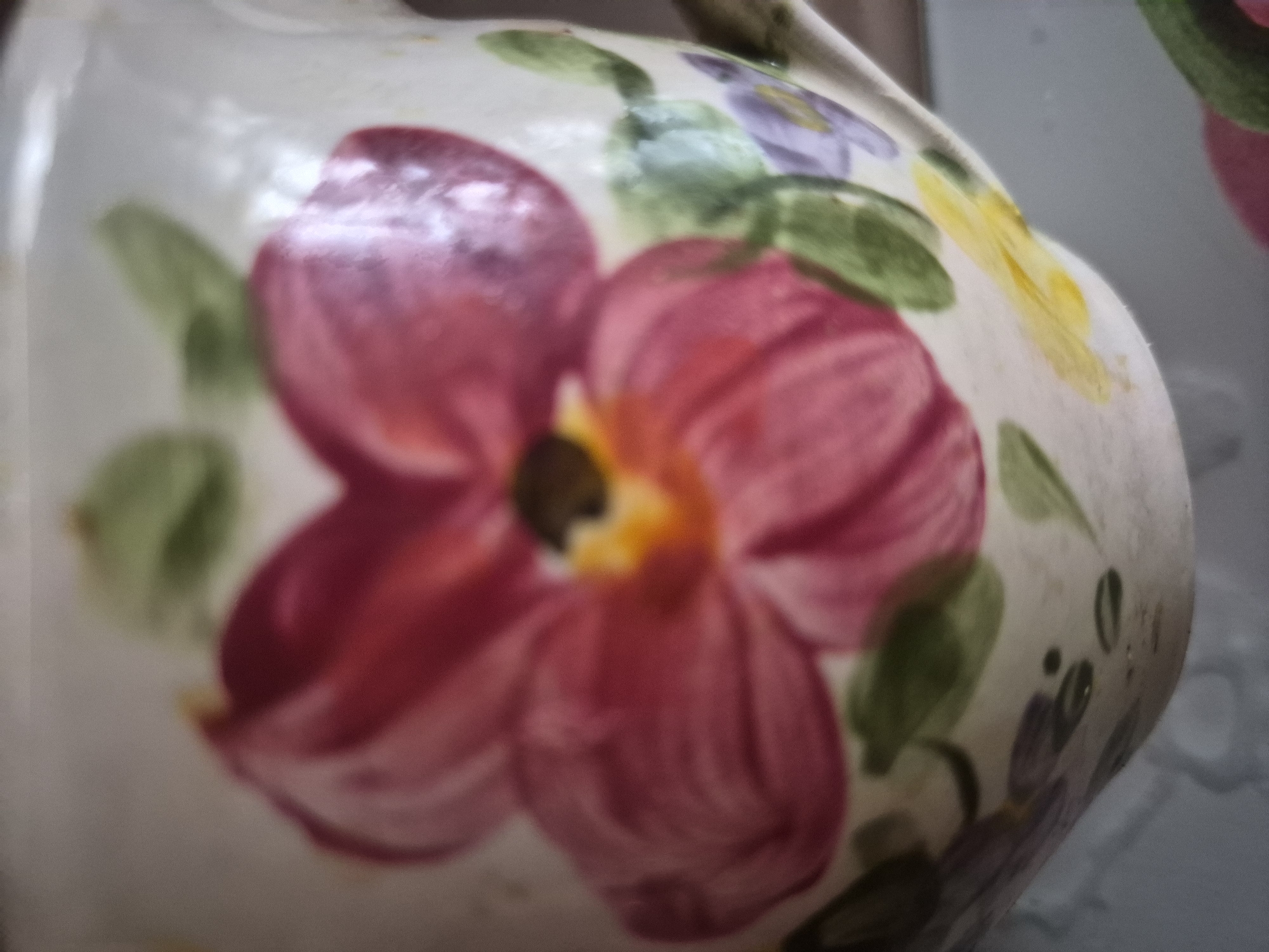 This close-up image features a glazed ceramic object, likely a pitcher or jug, adorned with hand-painted floral motifs. The object's base color is an off-white or cream, providing a soft background for the colorful decorations.
The most prominent feature is a large flower in the center, depicted in vibrant shades of pink and deep red, with a warm yellow and orange center. Surrounding this main flower are several green leaves, along with smaller floral elements in purple and yellow visible towards the top and sides of the object. The brushstrokes used for the painting are somewhat visible, suggesting a hand-painted technique.
The image has a shallow depth of field, with the central pink flower appearing relatively sharper, while the areas further from the primary focal point are softly blurred, creating an artistic effect. The surface of the ceramic appears smooth and reflective. No people are present, and no specific activity is occurring. The background is entirely out of focus, offering no discernible details about the setting. There is no visible text on the object. The item is located in Yerevan, Armenia.