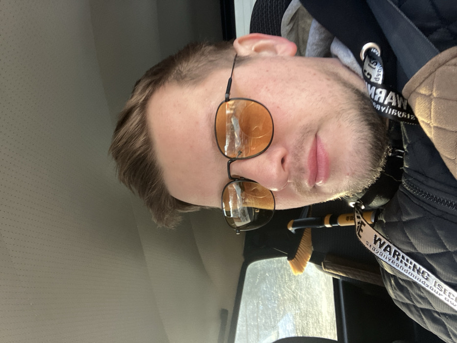 The scene depicts a light-skinned male individual seated in the front passenger compartment of a vehicle, located in Chaikovsky, Russia. The man has short brown hair and light facial stubble. He is wearing dark-rimmed sunglasses with orange-tinted lenses, which reflect ambient light. His attire consists of a dark, quilted jacket worn over a light grey hooded sweatshirt. A seatbelt is diagonally across his torso, secured with a beige, padded cover on the shoulder strap. Around his neck hangs a black lanyard with white text, partially legible as "WARNING IS SE..." and "MAY BE ALIVE AND MA...". He is looking directly towards the camera. The vehicle's interior features a light-colored, textured headliner. Through a rear window, a bright, illuminated outdoor environment is visible. Positioned behind the individual, within the vehicle, is a yellow broom with light bristles and a black and yellow striped handle, alongside an additional dark, elongated object.