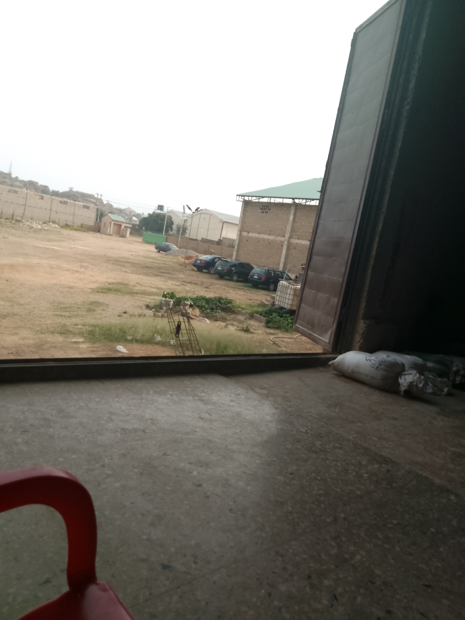 The image presents a static outdoor scene viewed from within a building, likely through a large open doorway or window. In the immediate foreground, a grey, speckled concrete floor is visible, with a partial red plastic chair in the bottom left corner. To the bottom right, two white sacks or bags rest on the floor, near a dark frame or threshold. Visible text on one bag includes "CORN FLOUR" and "NET WT. 50 KGS".
Beyond the opening, a large, unpaved area of dirt and dry, light green grass dominates the mid-ground. A dark horizontal bar or frame, possibly part of the window or door structure, spans the bottom section of the exterior view. Several dark-colored cars, appearing to be SUVs, are parked in a line to the right. To the left of the parked cars, a smaller structure with a light green roof stands. Further back, a long concrete wall runs horizontally, and behind it, hills or rocky formations are visible in the distance. Another, larger building with a green roof is seen to the right. A metal frame structure is visible in the grassy area.
The sky is overcast and hazy, indicating daytime without direct sunlight, possibly an early morning, late afternoon, or very cloudy day. The overall environment suggests an industrial or semi-developed area, consistent with the provided location context of Jos, Nigeria. No people or active events are depicted.