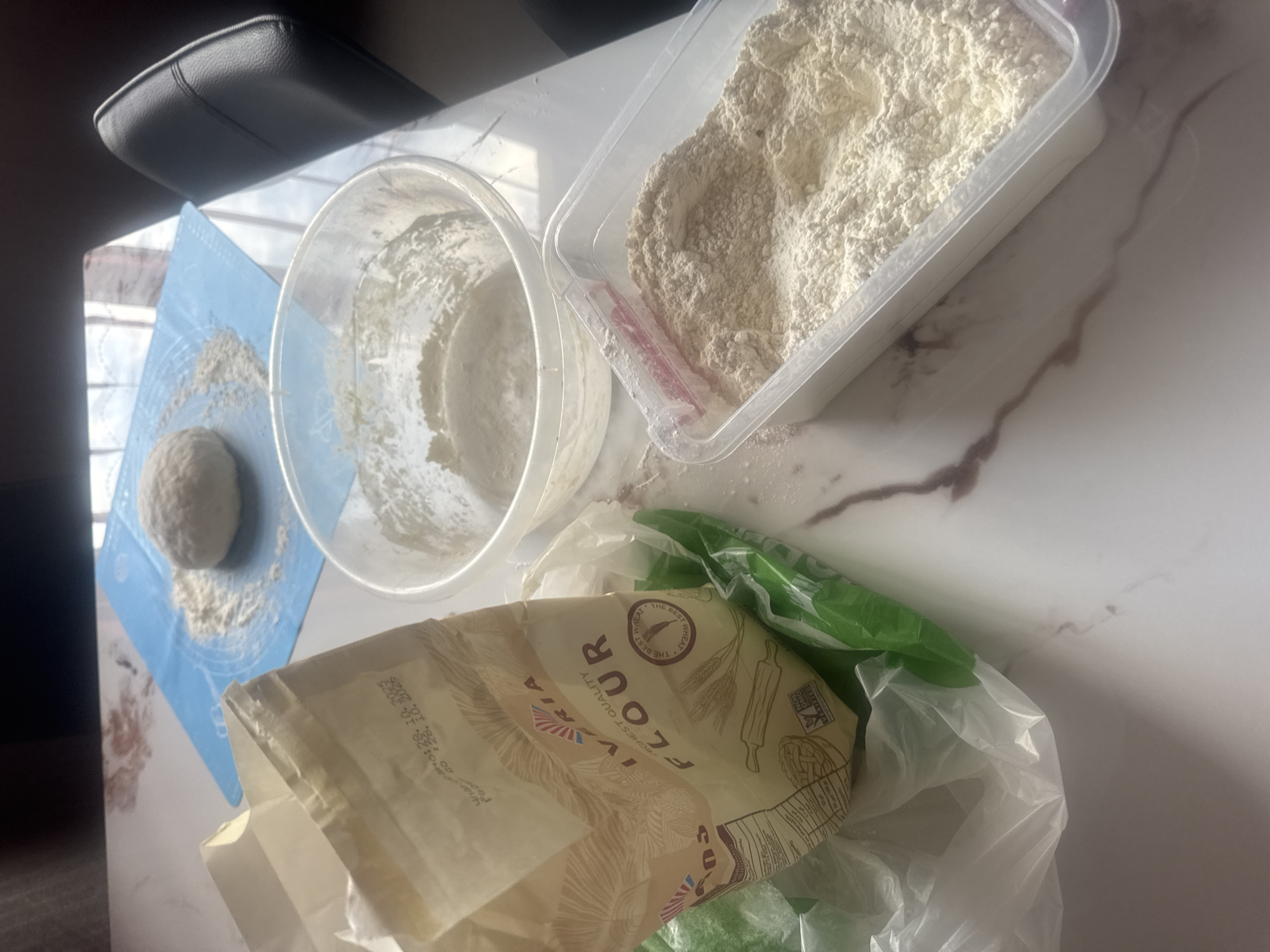 The image displays a bright, top-down view of a domestic baking or dough preparation scene on a white marbled countertop.
The primary subjects are various forms of flour and dough. In the upper right, a rectangular clear plastic container is filled with white flour. To its left, a clear plastic mixing bowl, coated with flour residue, is visible. Further left, resting on a light blue silicone pastry mat, is a small, round ball of dough, surrounded by scattered flour. The mat itself has faint circular markings and measurements.
Towards the bottom right, a large paper bag of flour, partially contained within a green plastic bag and a clear plastic bag, is prominent. The visible text on the paper flour bag includes "IVIRIA FLOUR," "HIGHEST QUALITY," and "THE BEST WHEAT." It also features an emblem of wheat and a rolling pin icon. A best-before date "2023.10.28" is partially legible on the bag.
The scene is well-lit, suggesting daytime, with strong light coming from the left, creating subtle shadows. In the upper left corner, a portion of a black upholstered armrest or chair is visible. The overall impression is one of an active kitchen space dedicated to baking, likely in the midst of making bread or other flour-based products.