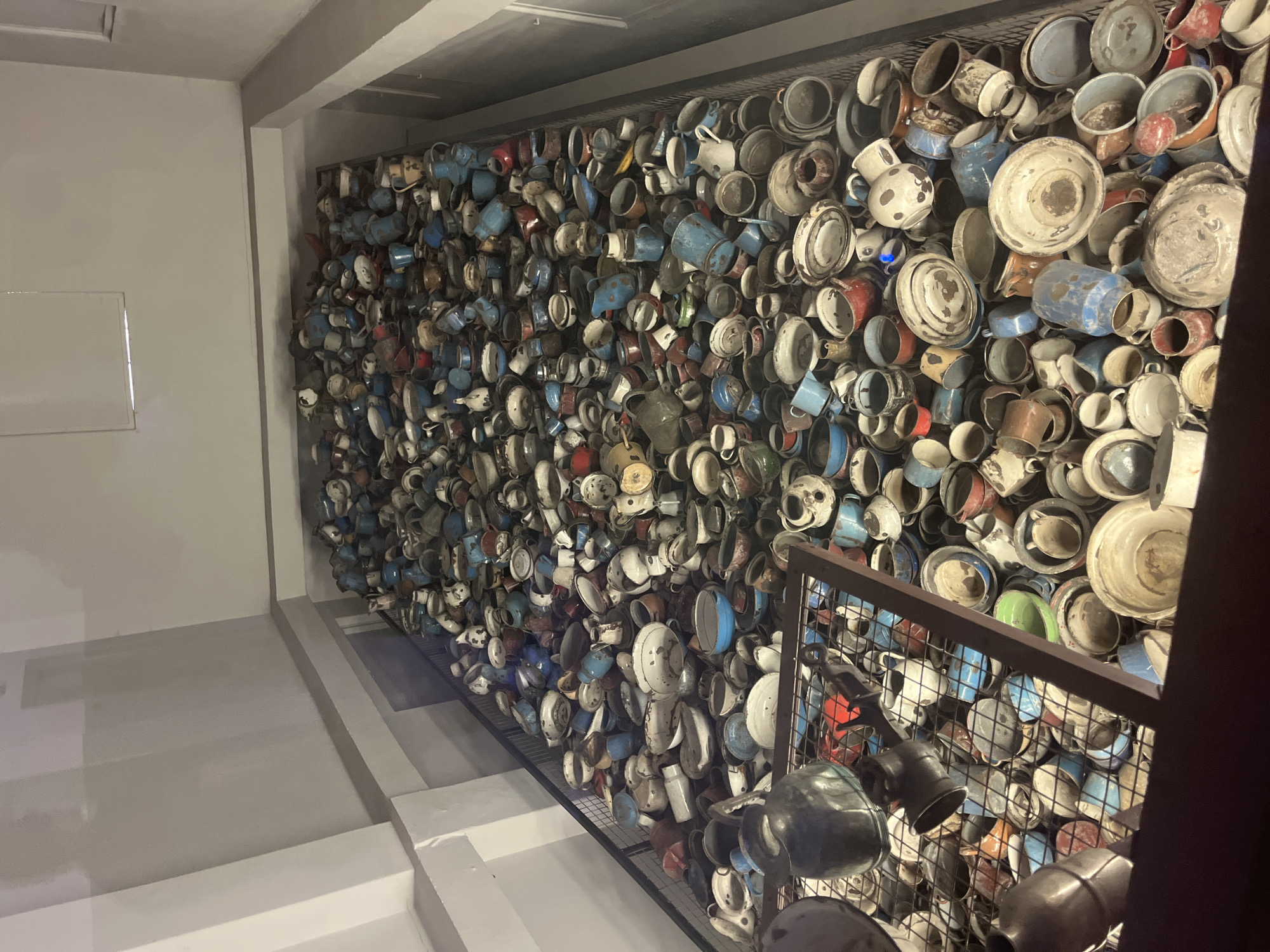 The image depicts a large collection of kitchenware, primarily pots, pans, and cups, piled high in a room. The objects are made of various materials, including metal and enamel, and are in shades of blue, white, red, and grey. Some items appear to be dented or worn. The room itself is utilitarian, with plain walls and a slatted floor or platform. This scene is located at the Auschwitz-Birkenau State Museum in Oświęcim, Poland, and represents a collection of belongings confiscated from victims of the Holocaust.