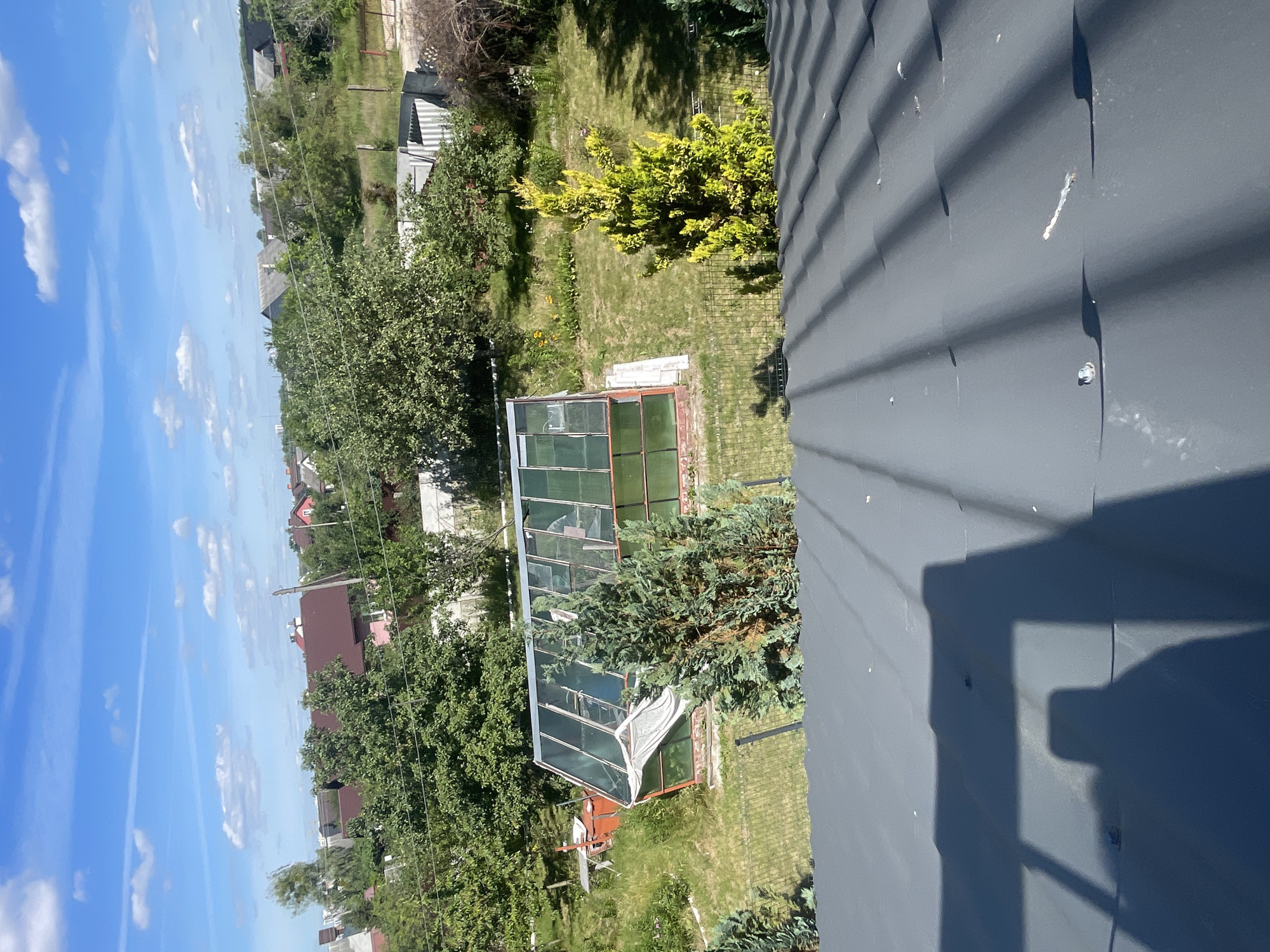 This is an aerial, high-angle photograph of a residential backyard in Мухавецкі сельскі Савет, Belarus, taken on a clear day with a bright blue sky and scattered white clouds. The foreground is dominated by the dark gray, corrugated metal roof of a building, with shadows cast by an unseen object, possibly the photographer.
In the mid-ground, a green and glass greenhouse with a red frame is visible. A white tarp or sheet is draped over one side of the greenhouse. Adjacent to the greenhouse, a bright orange structure, possibly a water tank or barrel, sits on a grassy slope. Several evergreen trees, one with yellow-green foliage, are scattered throughout the yard. A black metal fence encloses the garden area.
The background shows a suburban landscape with several houses featuring red and gray roofs, surrounded by lush green trees and vegetation. Power lines are visible crisscrossing the sky. The overall impression is a well-maintained, rural or suburban property on a sunny day.