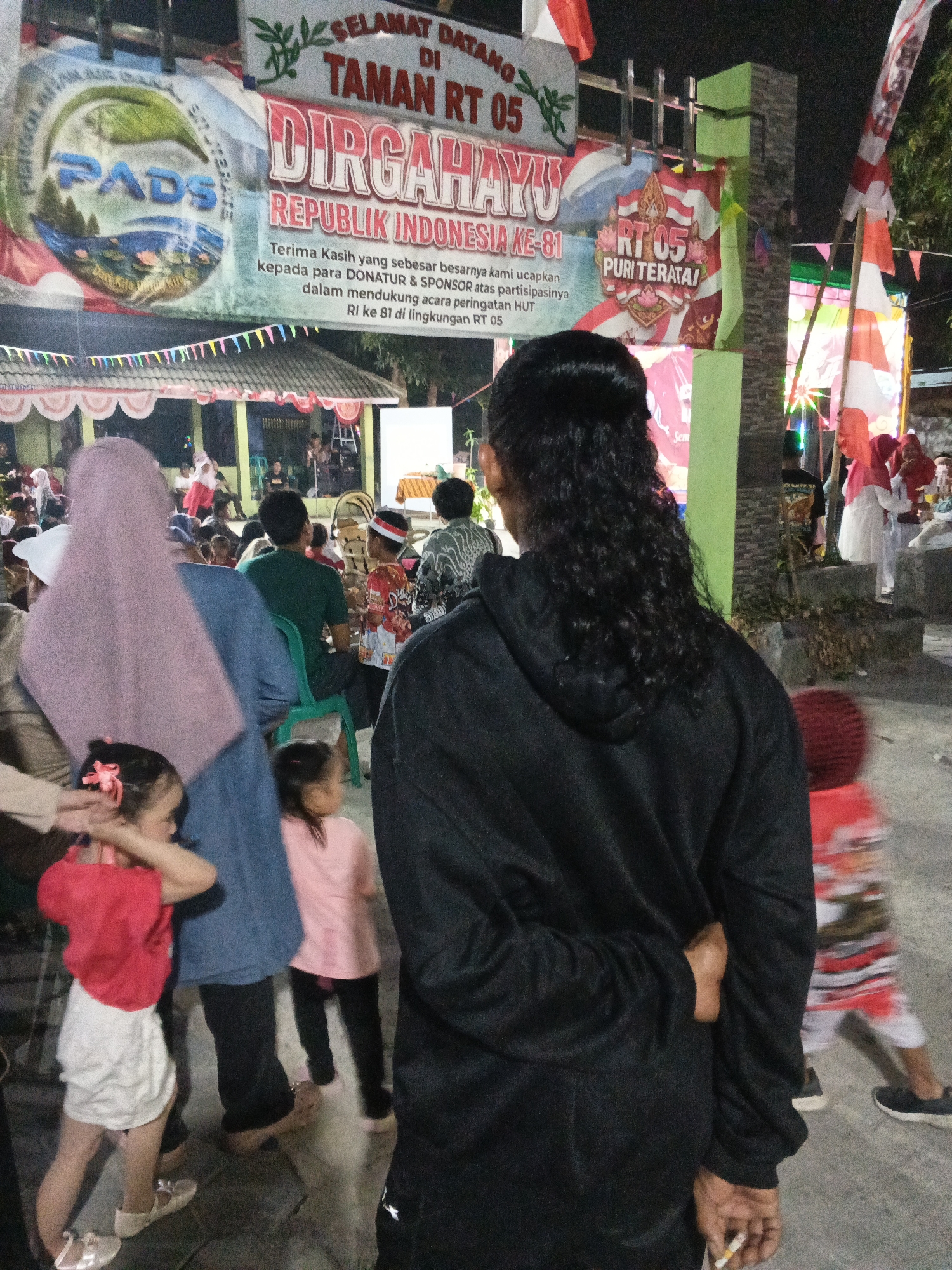 The image depicts an outdoor evening scene, likely a community event, in the city of Cikande, Indonesia. In the foreground, a person wearing a black hoodie stands with their back to the camera, holding a cigarette in their right hand. To the left, a young girl in a red shirt and white shorts, and another child in a pink shirt, are visible. In the background, a banner with Indonesian text, including "SELAMAT DATANG DI TAMAN RT 05 DIRGAHAYU REPUBLIK INDONESIA-81," suggests a celebration related to Indonesian Independence Day. Several people are gathered in front of a building adorned with Indonesian flags. The environment appears to be a public gathering space with makeshift decorations and lighting.