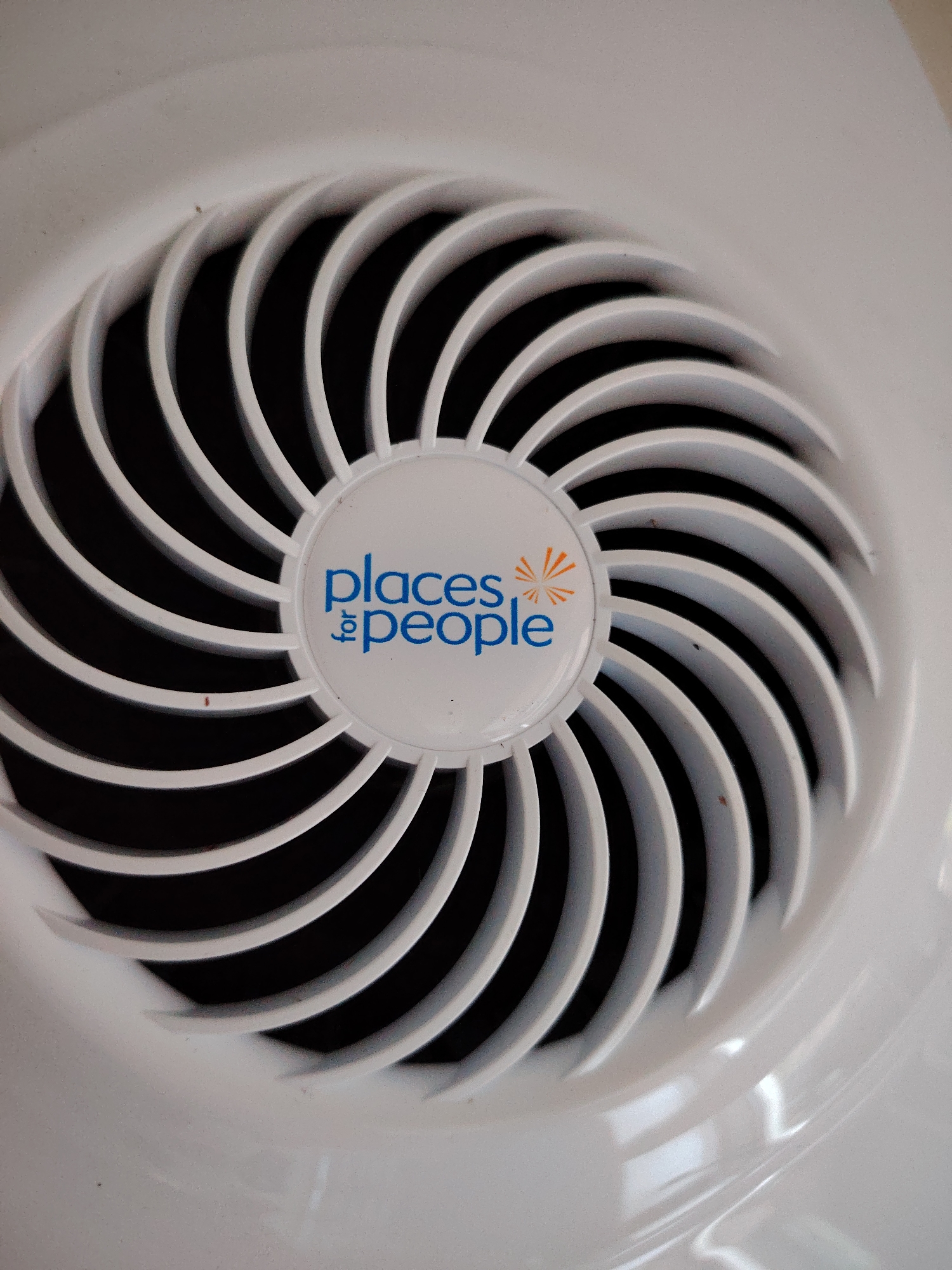 The image is a close-up, overhead shot of a white plastic fan grille. The grille has a swirling pattern of curved slats. In the center of the grille is a circular emblem with blue text that reads "places for people". The text is accompanied by an orange, stylized sunburst. The background behind the grille appears dark, suggesting the fan is in operation or the interior of the device is dark. There are some minor imperfections and dust specks visible on the grille. The image does not provide any information to determine its location in the city of Huntingdonshire, United Kingdom, nor does it show any people, actions, or interactions.