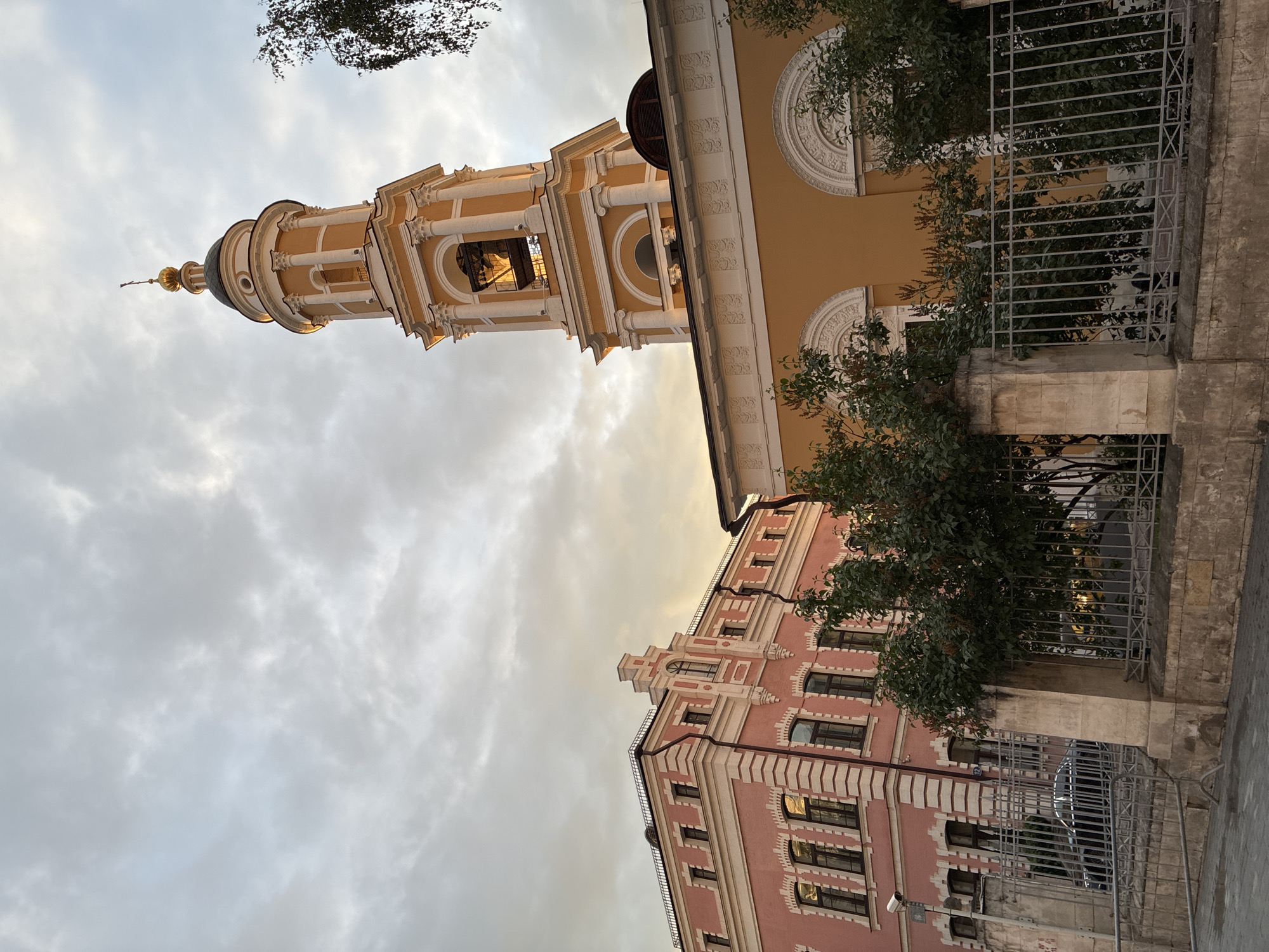 The image shows a view of buildings and the sky in Moscow, Russia. On the right is a yellow building with white architectural details, including round decorative elements and a bell tower with a golden dome and cross at the top. In front of the yellow building is a gray metal fence and green foliage. To the lower left, there is a pink building with white trim and arched windows. The sky is overcast with gray and white clouds. The perspective of the image is angled upward, showcasing the height of the buildings against the sky.