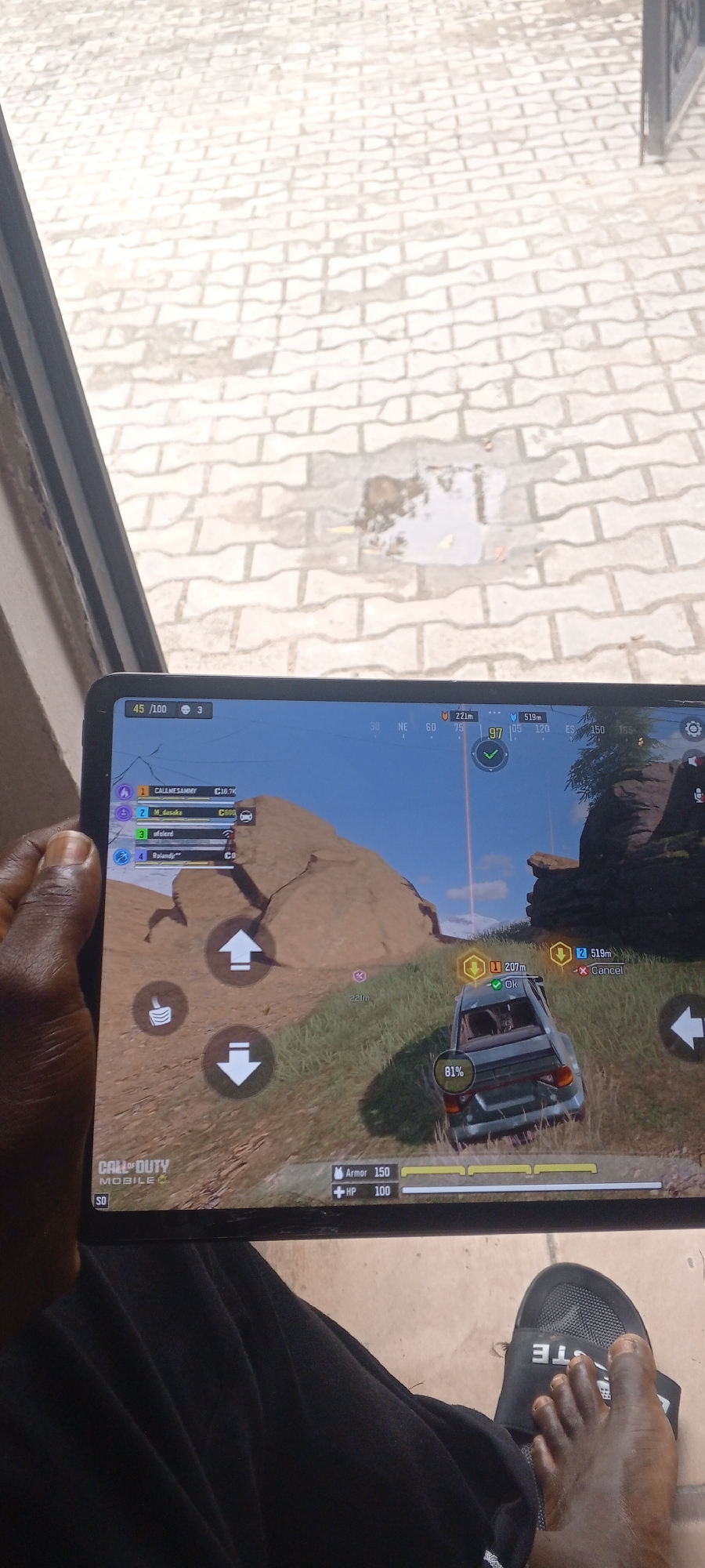 The image shows a person playing a mobile game, likely Call of Duty Mobile, on a tablet. The tablet is held by the person's left hand, with their feet and legs visible in the foreground. The game screen displays a battle royale scenario with a car in the middle of the frame, surrounded by a rocky, grassy terrain under a bright blue sky. The player's character is in the car, indicated by the "81%" health bar. On-screen indicators show player names like "CALLMESAMMY" and distances to other players, along with game controls like directional arrows. The setting appears to be outdoors, with a paved courtyard visible in the background, suggesting a daytime activity. The person is wearing black pants and black slides with the brand name "LACOSTE" visible on the right foot. The scene implies a casual activity, likely taking place in a residential or public outdoor space in Ilado, Nigeria, given the provided location context.