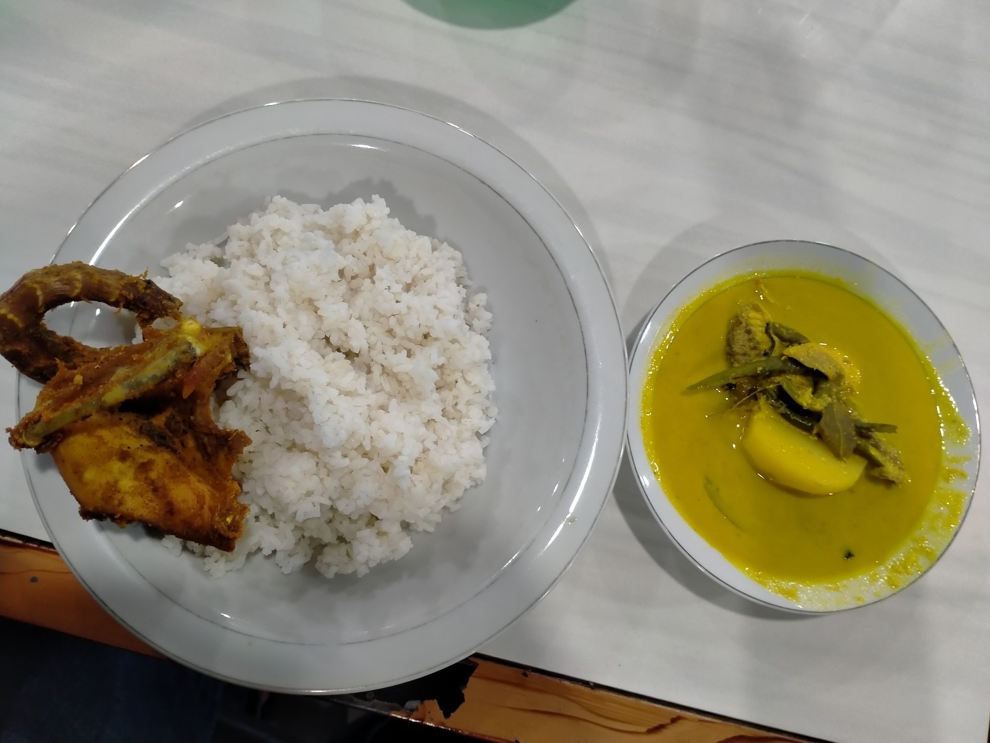 The image depicts a meal laid out on a white surface, likely a table. On the left, a white plate contains a mound of white rice and a piece of fried chicken or fish. To the right of that, in a separate white bowl, is a yellow-colored soup or stew, with visible pieces of potato and green vegetables within. The food appears to be Indonesian cuisine. There is no information in the image to confirm the location.