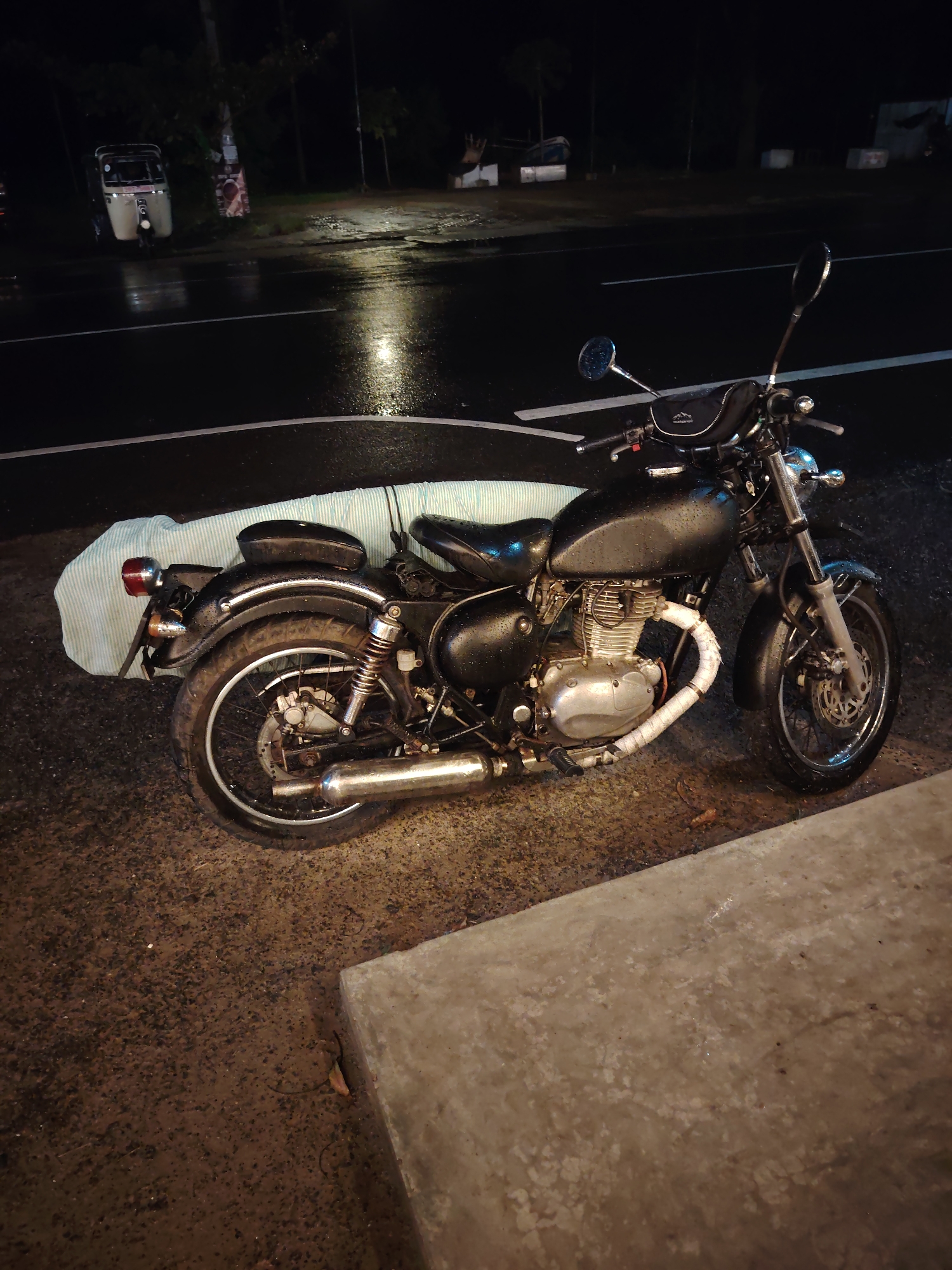 The scene depicts a static, nighttime environment in the city of Unknown, Unknown, following or during a rain event. A black, classic-style motorcycle, possibly a Royal Enfield, is parked on a damp, textured ground surface in the foreground, adjacent to a concrete curb on the right. The motorcycle's engine has a silver finish, and its exhaust header is wrapped in white material. Water droplets are visible on the motorcycle's tank, seat, and other surfaces. A black pouch with "MOUNTAIN RIDE" text is attached to the handlebars, and a light blue and white striped fabric object is strapped to the rear seat.
In the mid-ground, a dark, wet asphalt road with reflective puddles and white painted markings stretches horizontally across the frame. To the left of the road, a light-colored auto-rickshaw is stationary near dark foliage and a pole bearing a poster. The background is predominantly dark, with indistinct shapes of trees and possibly distant structures under minimal illumination. No individuals are visible in the image.