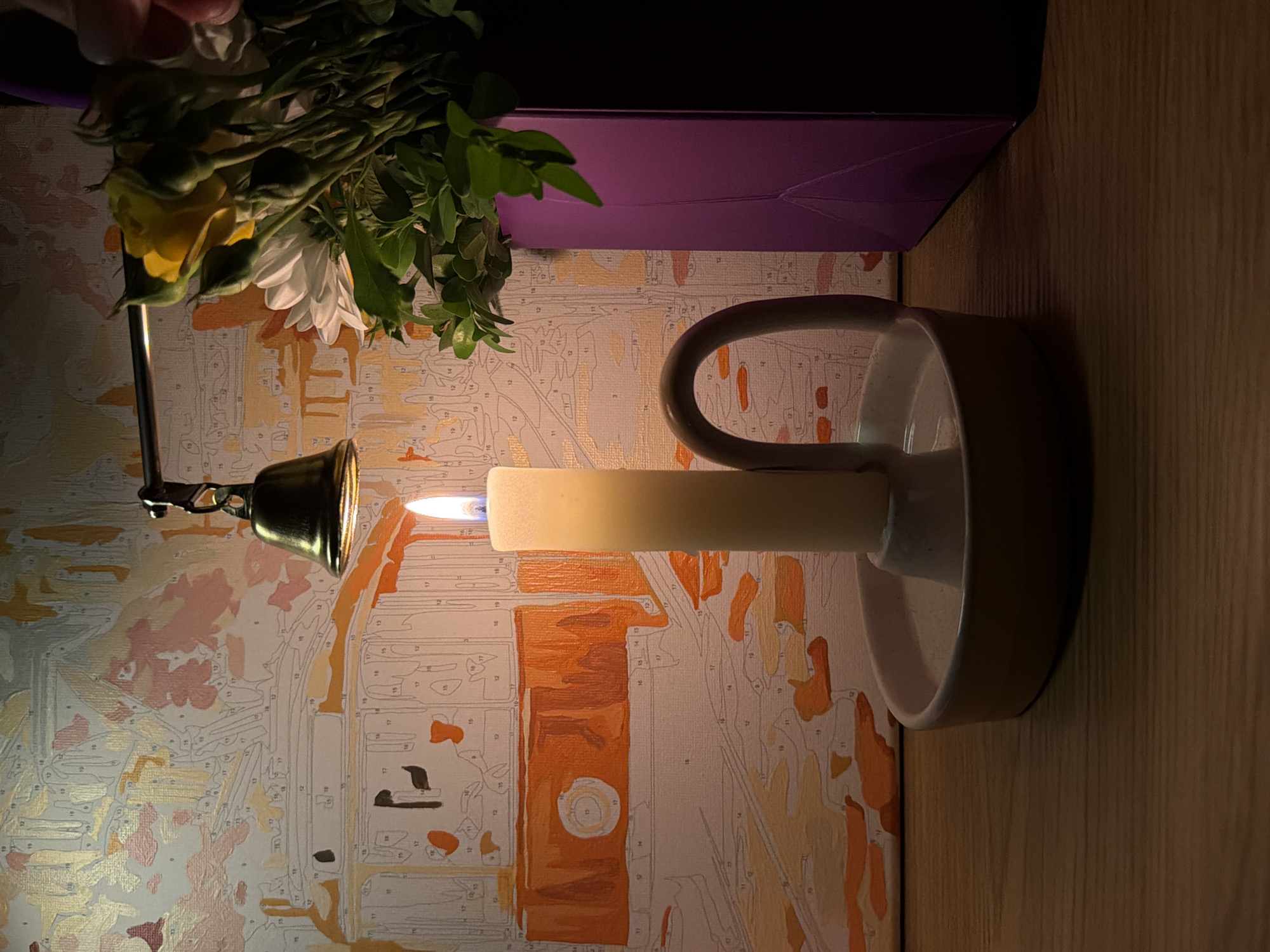 A dimly lit image shows a lit candle in a white candlestick holder, with a brass bell positioned next to the flame and a bouquet of flowers and a purple object in the background. The candle flame is small and bright. A paint-by-numbers canvas is visible behind the candle and flowers, displaying numbered sections outlined in gray and filled with various colors, including shades of orange, white, and beige. A wooden surface is visible on the right side of the image, next to the candle holder. The location of this image is Unknown, Unknown.