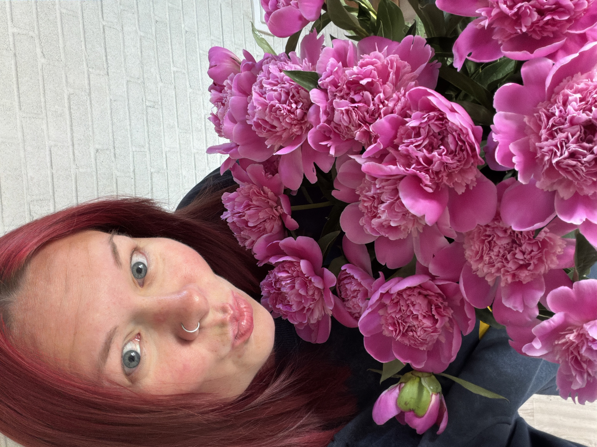 A selfie of a woman with red hair and a silver nose ring, holding a bouquet of bright pink peonies. The background appears to be a white brick wall. Location: Unknown, Unknown.
