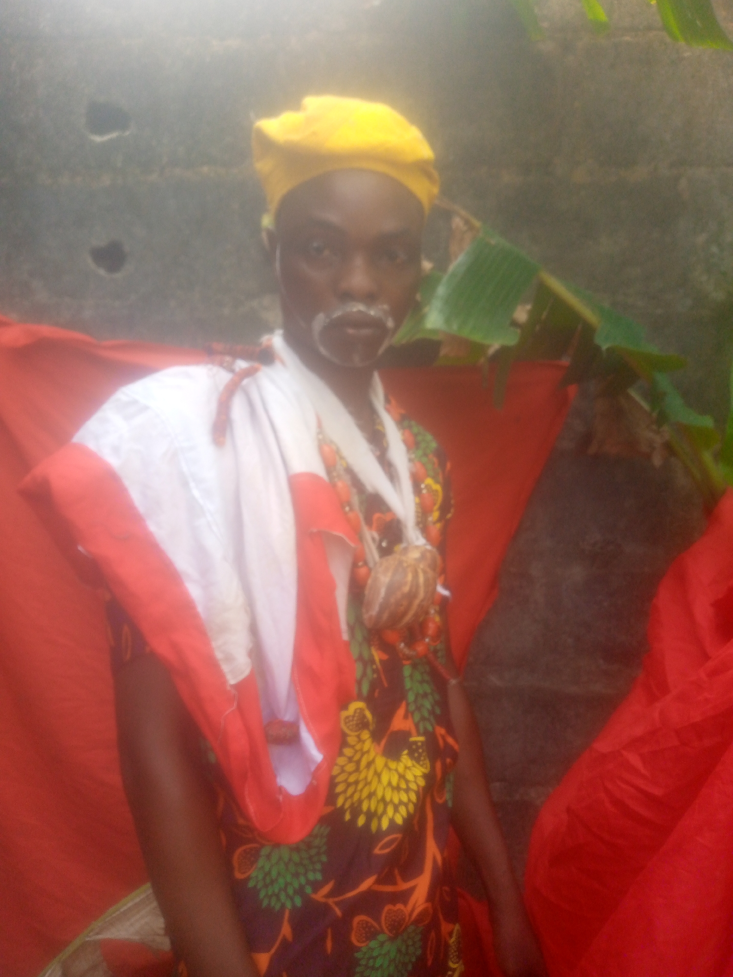 A full-shot shows a man wearing traditional attire, standing in front of a red cloth backdrop in an outdoor setting. The man is dark-skinned, with white makeup around his mouth, resembling a beard. He wears a yellow cap on his head, and a patterned shirt featuring yellow, green, and brown designs. He also wears a white cloth with red edges draped over his shoulder and a beaded necklace with a large shell pendant. Behind the man is a cement block wall with green foliage. A red cloth is also draped behind him. The location appears to be somewhere in Unknown, Unknown.