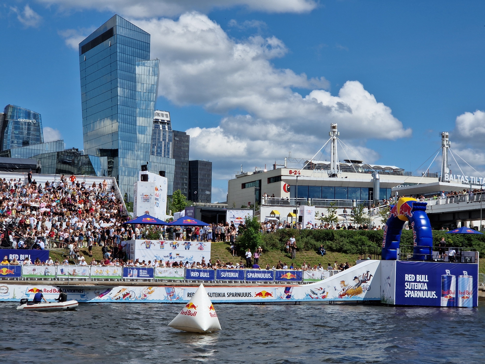 The image depicts a Red Bull event taking place on a river in Vilnius, Lithuania. In the background, modern skyscrapers, including the "Eastnine" building, and other urban structures rise against a sky with scattered white clouds. In the foreground, a ramp structure adorned with Red Bull branding extends over the water. Spectators are visible in tiered seating to the left and along the riverbank. A small inflatable boat with two individuals is present on the water, and a white, pyramid-shaped buoy with Red Bull logos floats nearby. The scene suggests an outdoor sporting event or competition, likely a stunt or water-based activity.