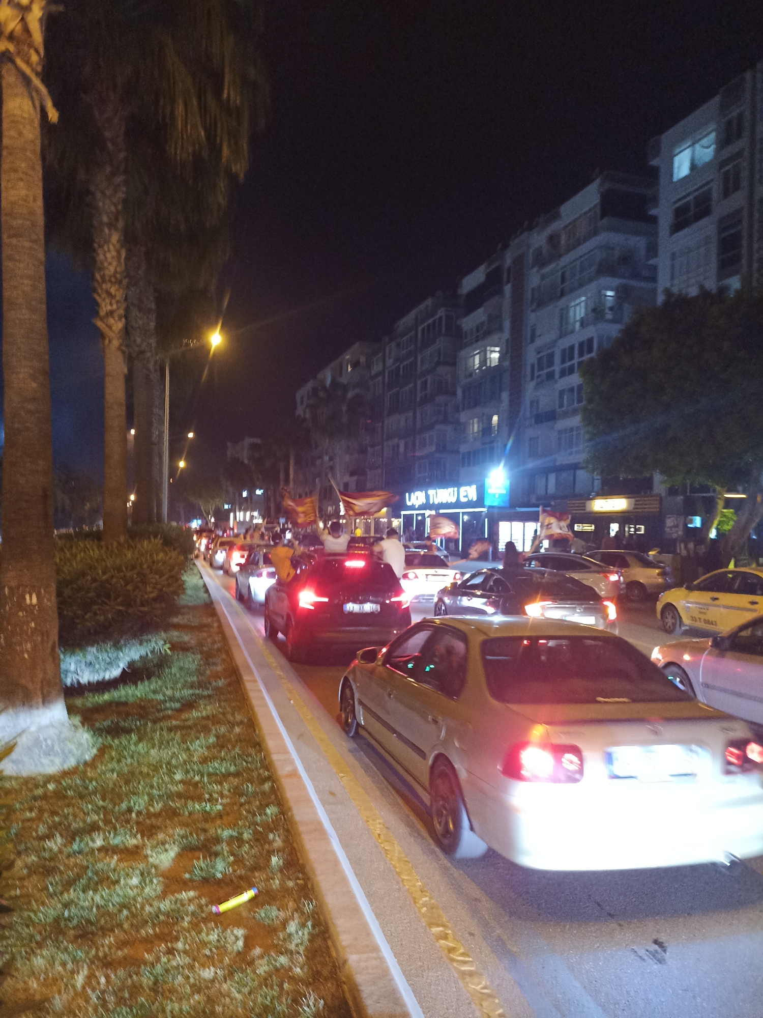 The image shows a nighttime street scene in Yenişehir, Turkey. A line of cars is stopped or moving slowly on a road lined with palm trees. Some people in the cars are waving flags. Buildings with lit windows are visible on the right side of the road, and a business with a sign in Turkish can be seen in the background. There are people standing in the area, possibly watching the procession of cars. The lighting is provided by streetlights and the headlights and taillights of the vehicles.