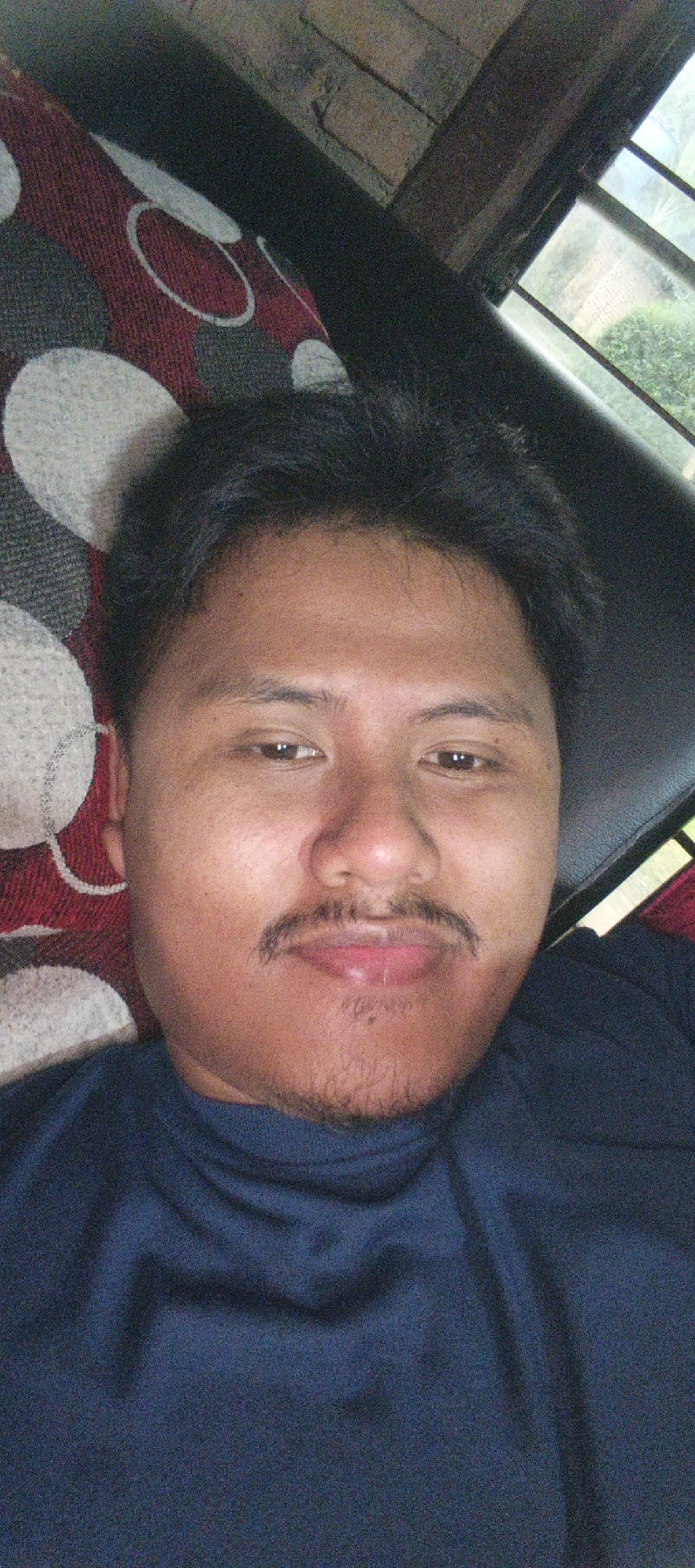 A close-up, slightly angled selfie of a young man is captured from the chest up. He has dark hair, brown eyes, and a mustache and light stubble. He is wearing a dark blue shirt. He is positioned against a background that includes a patterned fabric with circles in red, gray, and white, and a dark surface that could be furniture. To the right, a window with a view of greenery and a brick wall is visible. The lighting suggests an indoor setting, possibly during the daytime. The location in Kemuning, Indonesia is not discernible from the image itself.