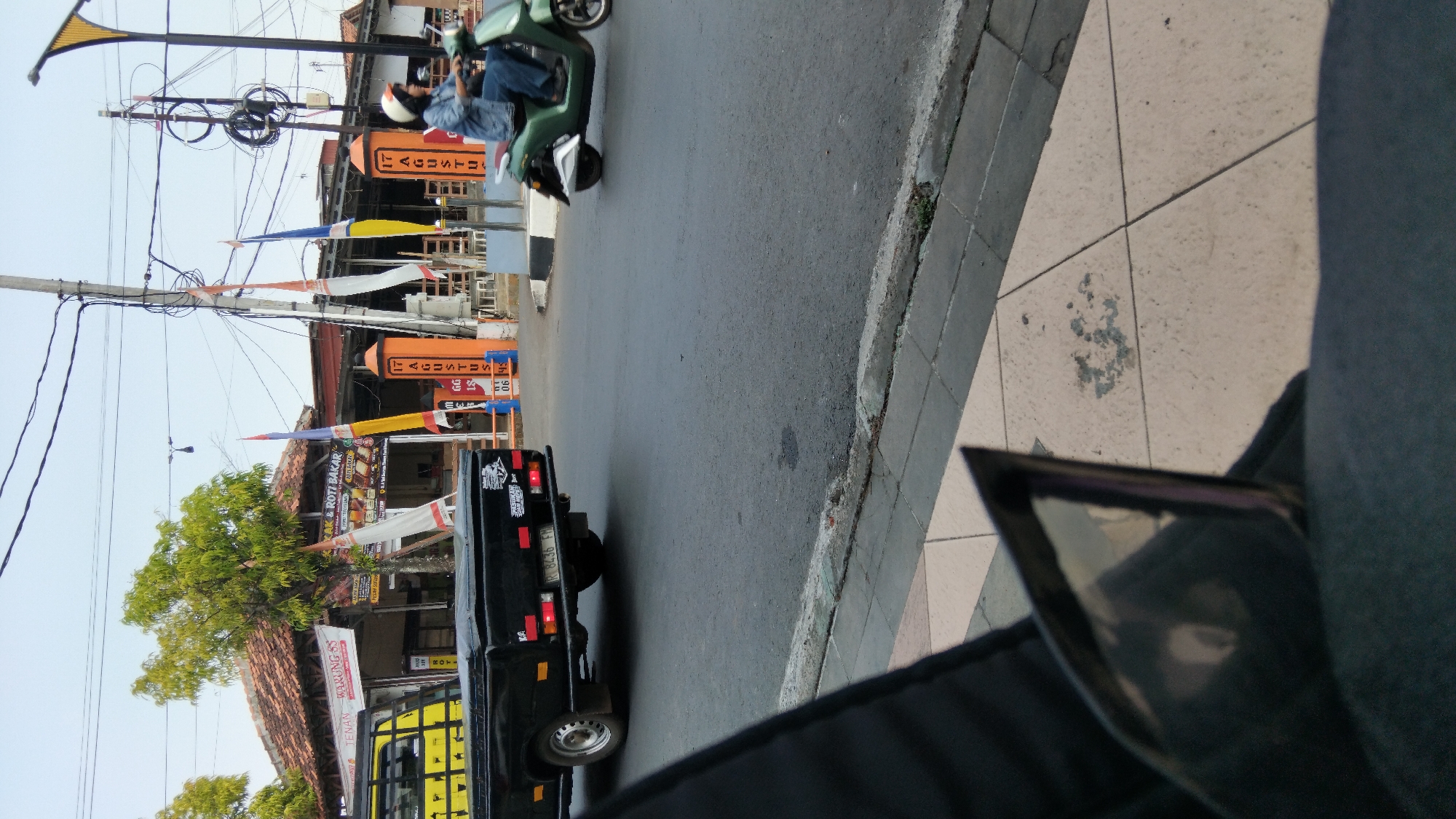 This is a street-level shot in Batang, Indonesia, likely on Jalan Yos Sudarso. A black pickup truck is parked on the left, its rear visible. In the middle ground, a person on a green scooter rides away from the camera. Behind them, a building with a tiled roof stands, and banners with Indonesian text and national flags are displayed, possibly indicating a local business or event. The foreground shows a sidewalk and a portion of a vehicle's interior.