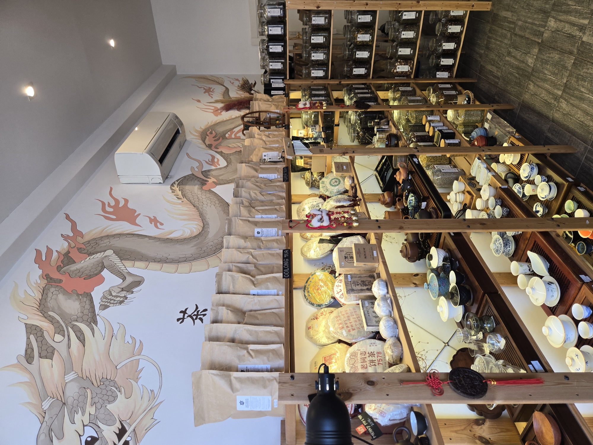 The image captures the interior of a tea shop in Budva, Montenegro. A prominent dragon mural adorns the wall, adding an oriental touch to the setting. Shelves filled with various tea products such as Oolong and GABA tea, teapots, and cups are arranged neatly, creating a well-stocked and visually appealing display. Jars filled with tea and tea leaves can also be seen on the upper shelves. Additionally, there's an air conditioning unit installed high on the wall. The presence of Chinese characters and oriental-themed decorations further highlight the shop's dedication to tea culture.