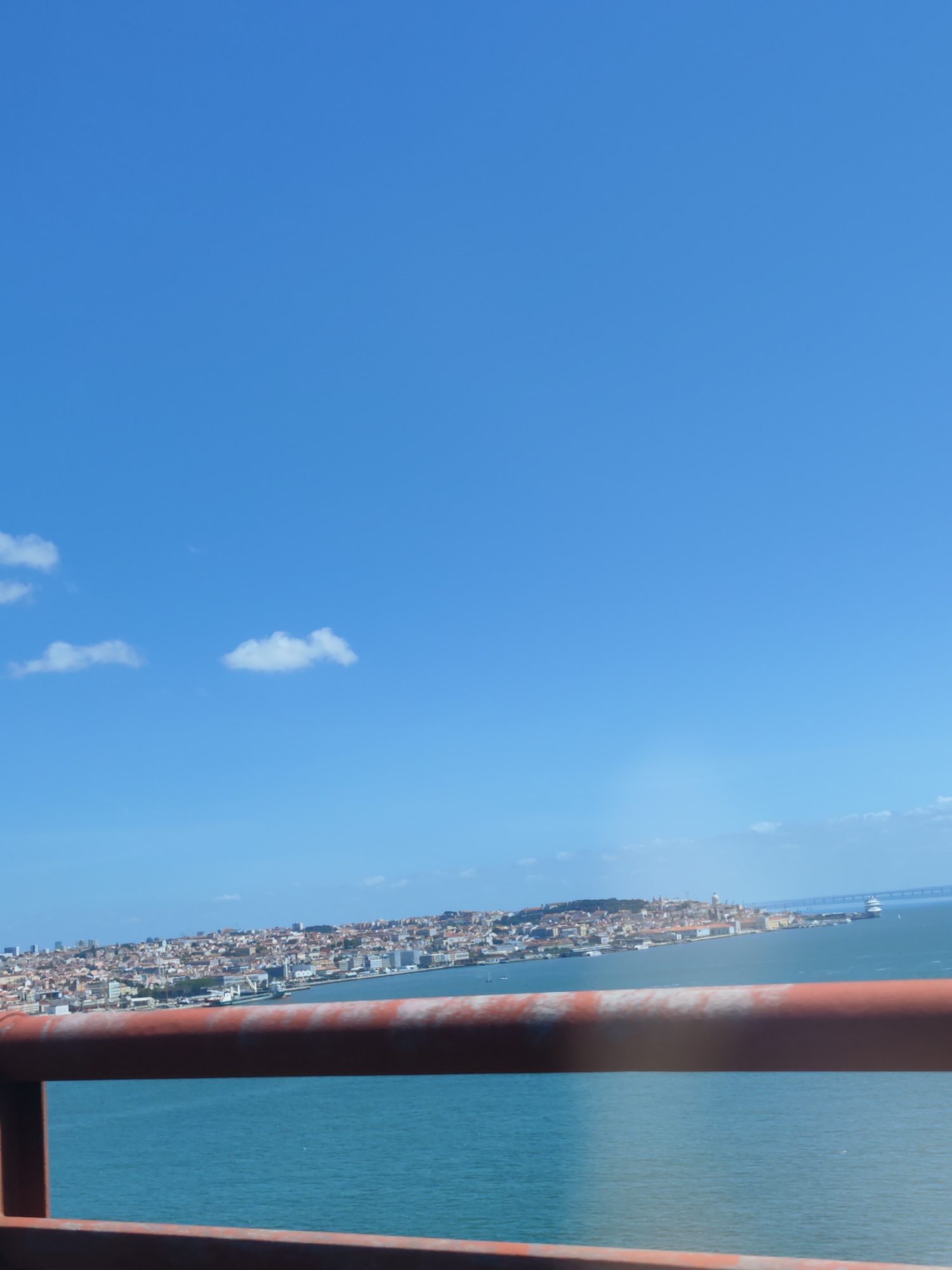 The image shows a view from a bridge in Lisbon, Portugal, overlooking the city. A rusty red metal railing is visible in the foreground. Beyond the railing is the Tagus River, and on the opposite bank is the cityscape of Lisbon. The sky is blue with a few scattered clouds.