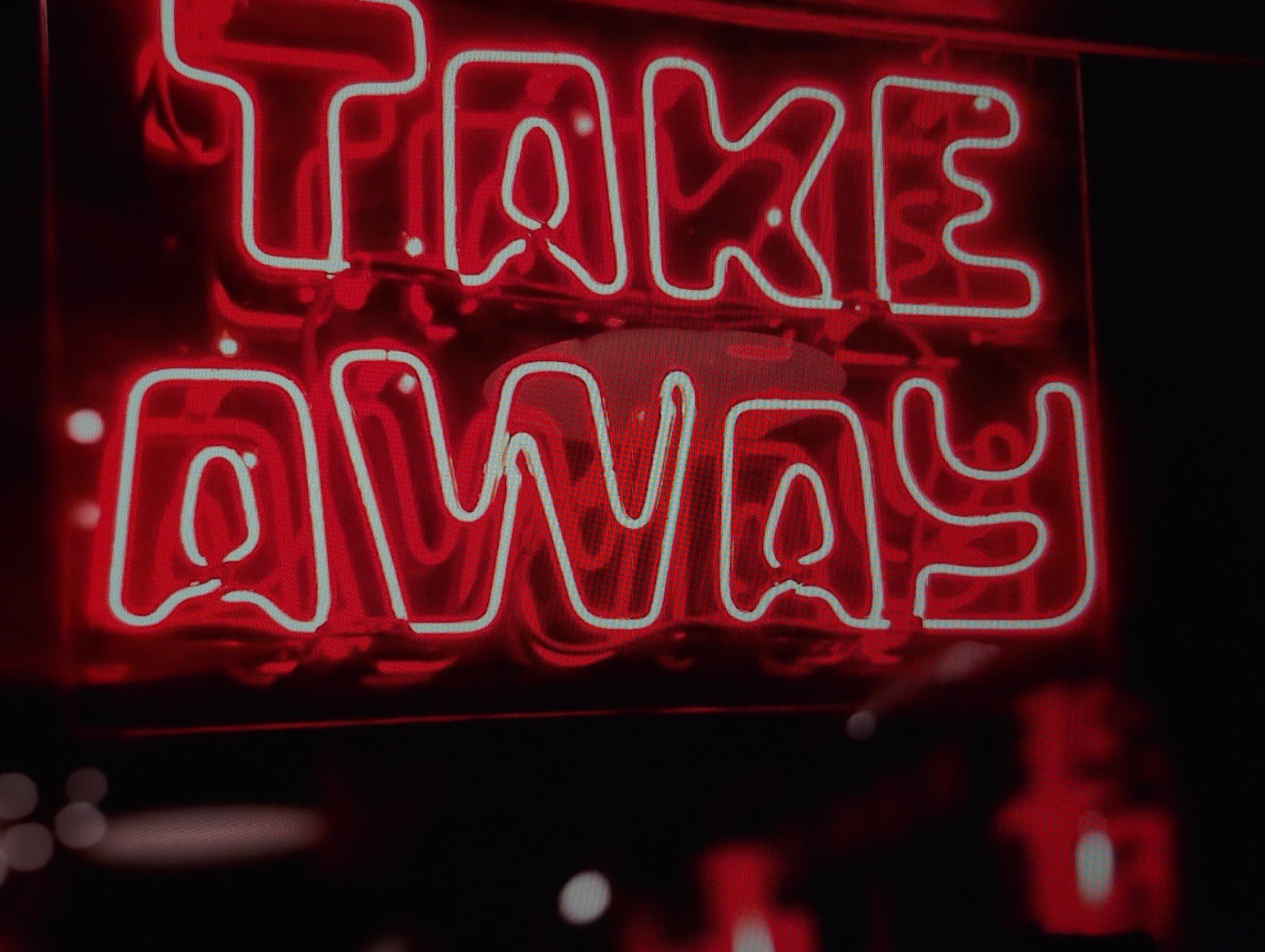 A close-up shot captures a red neon sign with a white outline spelling "TAKE AWAY." The background is black, with blurred red lights and white circles suggesting other light sources. The location in the city of Izhevsk, Russia is not apparent from the image.