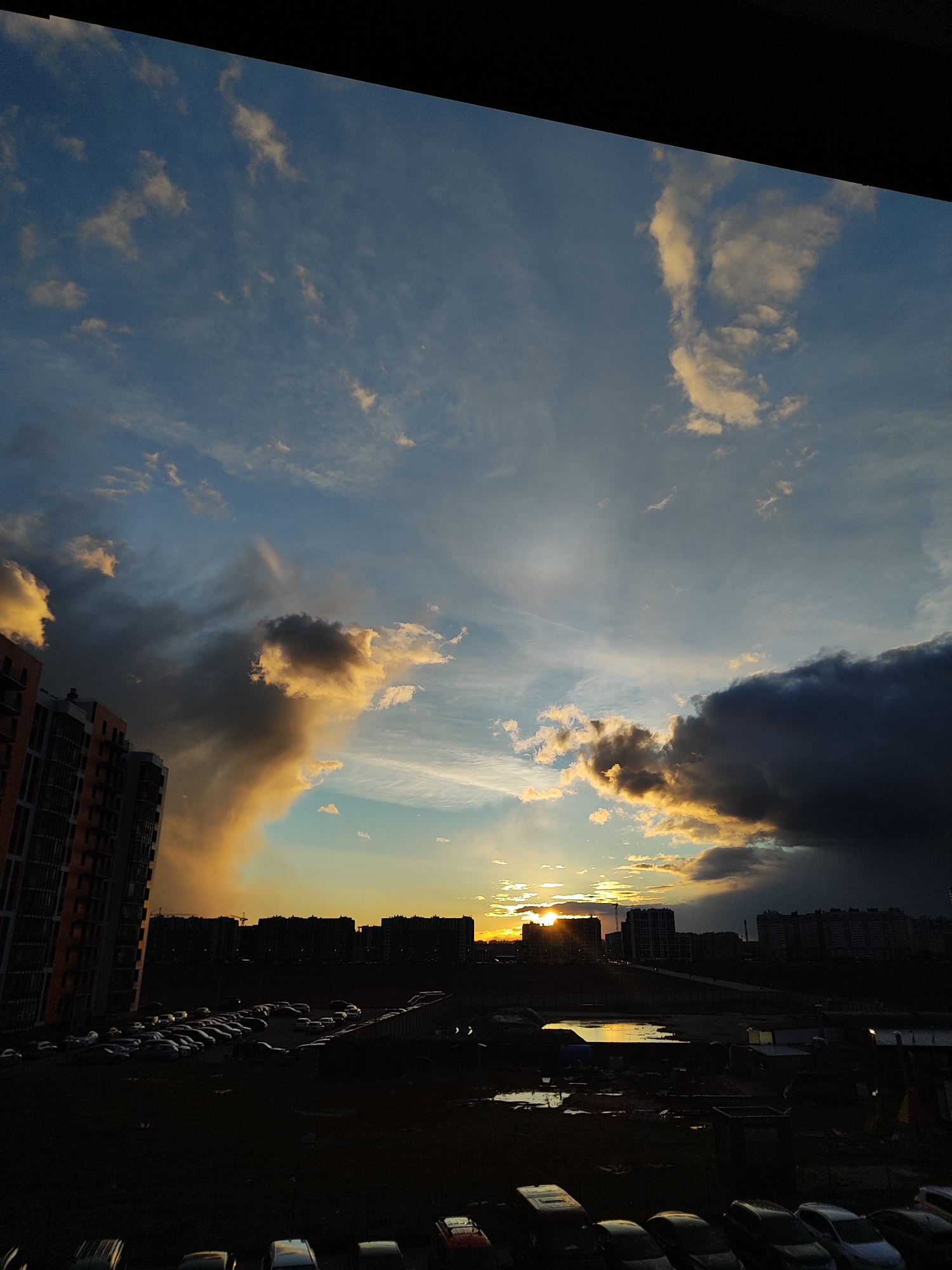 The image captures a dramatic sunset over an urban or suburban landscape, viewed from an elevated perspective, possibly a window, indicated by the dark frame at the top. The sky is a prominent feature, displaying a striking contrast between areas of clear blue, bright golden and orange clouds illuminated by the setting sun, and large, dark, heavy storm clouds on the left and right. The sun is visible as a bright, glowing disc just above the horizon, partially obscured by distant buildings and clouds, casting warm light across the mid-ground clouds.
In the foreground, the scene is heavily silhouetted and dark, revealing a sprawling parking lot filled with numerous cars. Beyond the parking lot, there's an area of uneven ground, possibly muddy or partially flooded, reflecting patches of the colorful sky. The middle ground and background are dominated by the dark silhouettes of numerous multi-story residential or commercial buildings, forming a city skyline. Some distant structures, possibly including a crane, are faintly visible against the horizon. No people are visible, and there is no discernible text. The time of day is clearly sunset, characterized by the intense, low-angle light and strong shadows, while the presence of dark clouds suggests dynamic weather, possibly after a rain shower or with an impending storm.