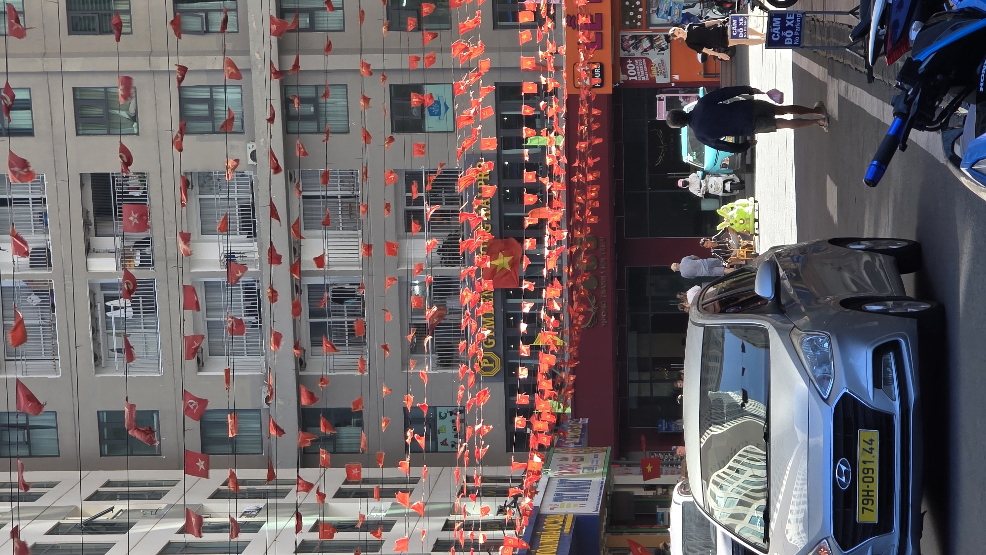The image depicts a street scene in Bac Nha Trang Ward, Vietnam, adorned with numerous Vietnamese flags strung between buildings. Pedestrians are walking on the sidewalk, and a gray Hyundai car is parked on the side of the road. Several motorcycles and scooters are also parked nearby. The foreground features the car's license plate, which reads "79H-091.44." A building with "PIGMEN" in large letters is also visible in the background. The sky is bright, suggesting a sunny day.