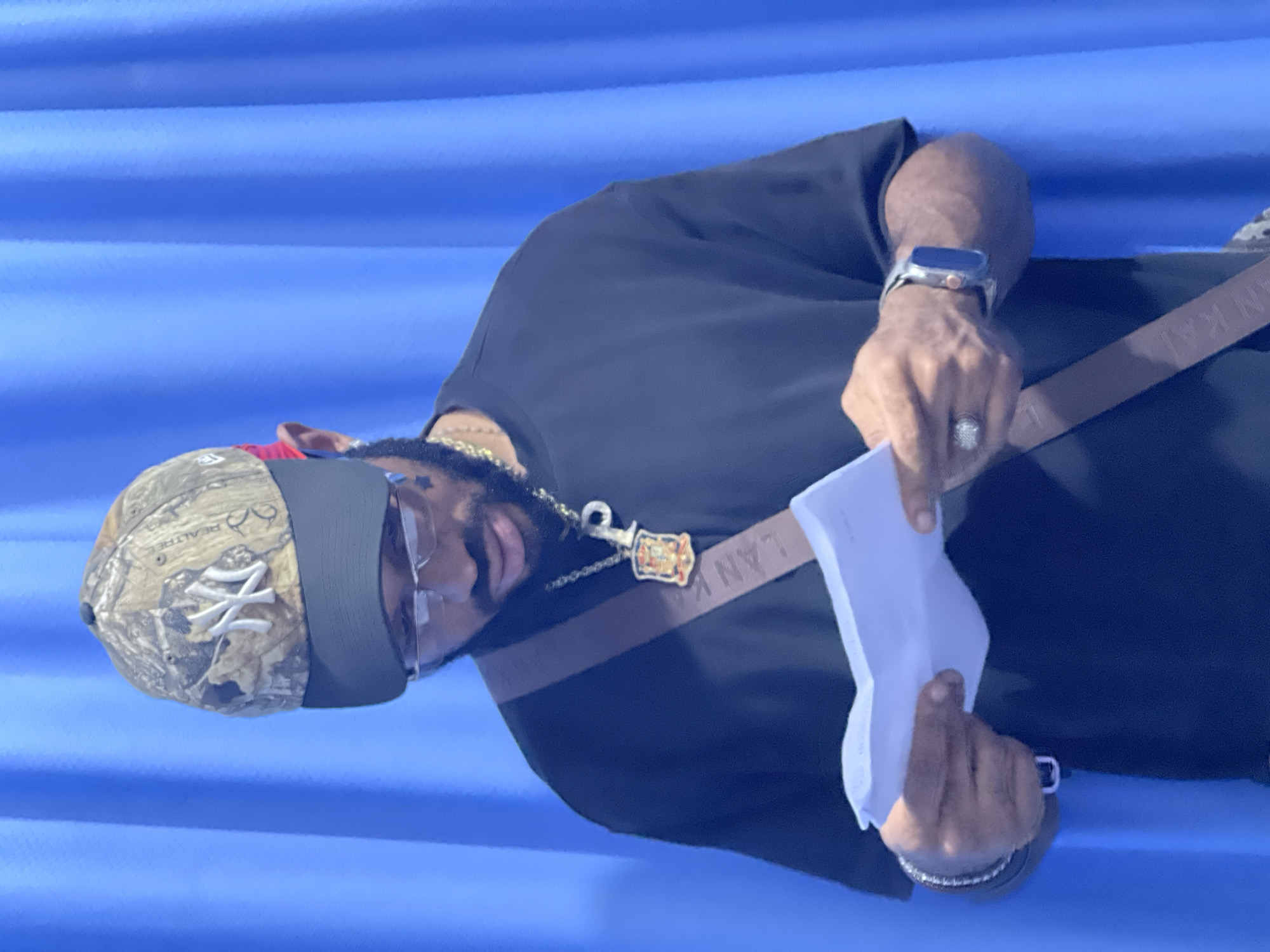 The image shows a Black man standing in front of a blue, pleated backdrop. He is wearing a camouflage baseball cap with a New York Yankees logo, glasses, a black t-shirt, a gold chain with a pendant, a silver watch, and several bracelets. He is holding a white piece of paper in his hands and appears to be reading it. The man has a beard and is wearing a ring on his left index finger. A brown strap, possibly from a bag, is visible across his chest. The lighting suggests an indoor setting, and the backdrop's uniformity indicates it might be a studio or a staged environment. The image does not contain any identifiable landmarks or clues to definitively place the location within Abuja, Nigeria, or any other specific city.