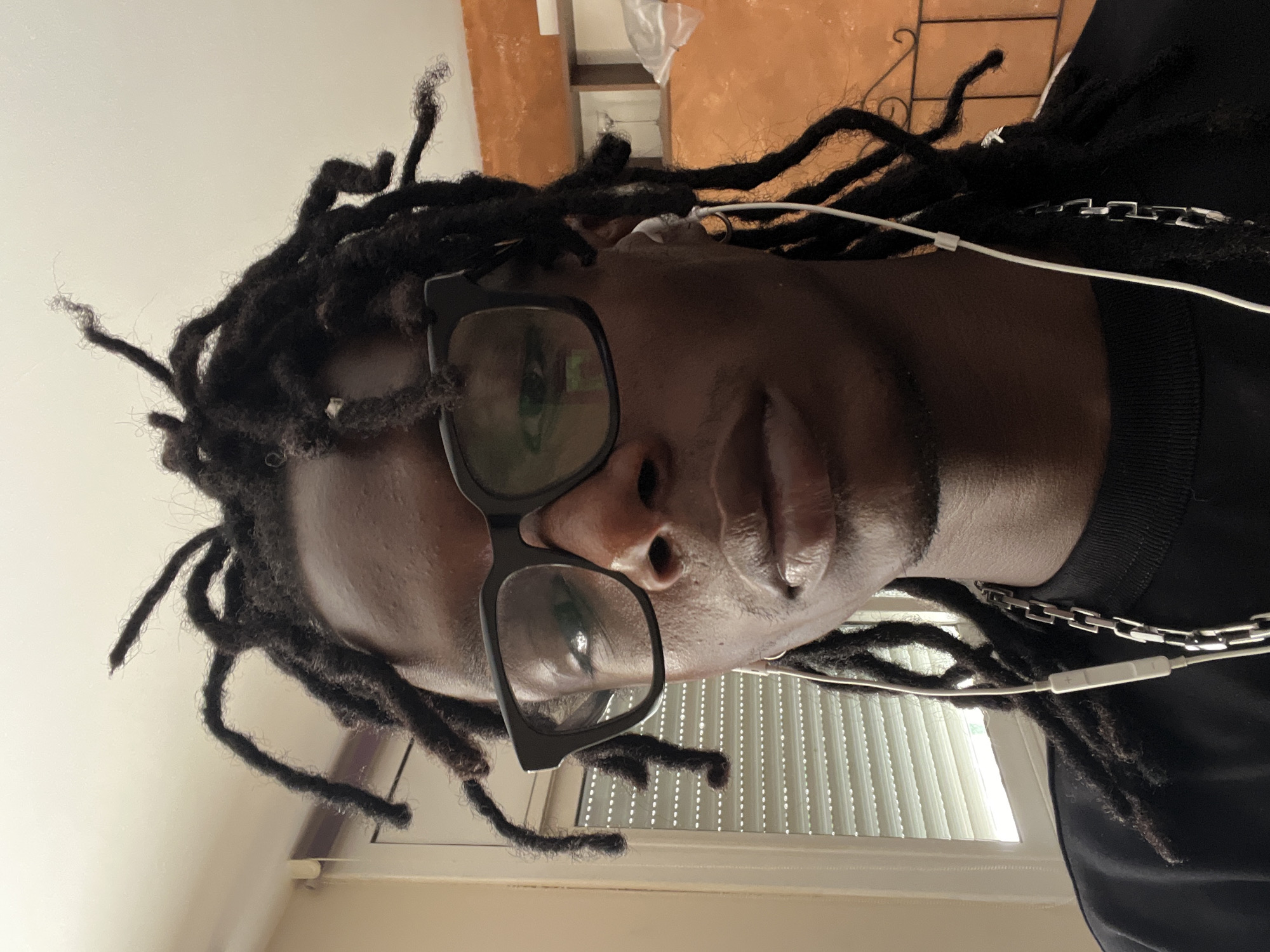 The image is a close-up, front-facing portrait of a young Black person with dreadlocks, wearing thick-rimmed black glasses and white earbuds. They are wearing a black t-shirt with a thick silver chain necklace. The background is an indoor setting with a window covered by blinds on the left and an orange wall with shelves on the right. The person is looking directly at the camera. The lighting appears to be natural, coming from the window. There is no indication of the location being Barcelona, Spain.