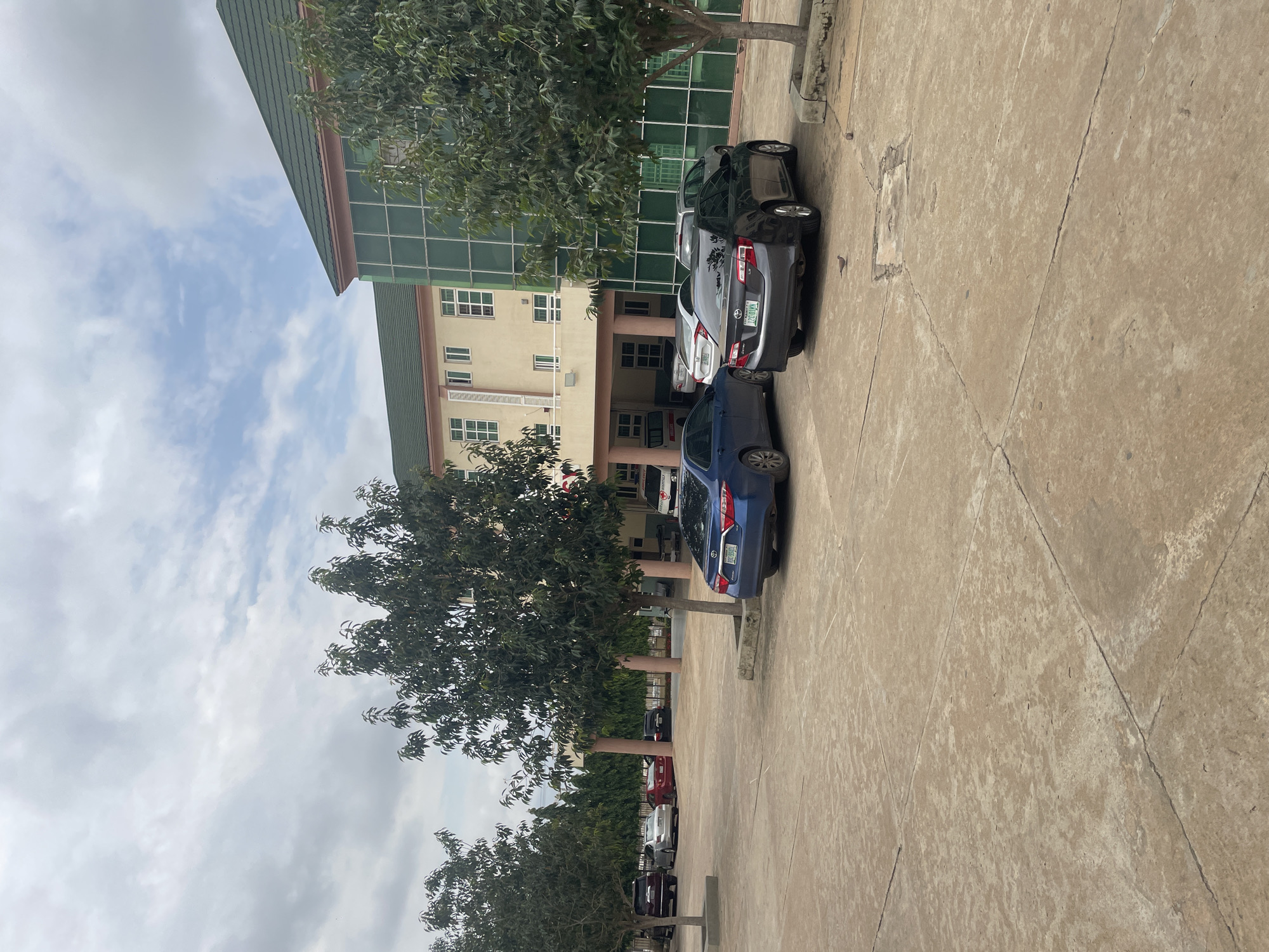 The image depicts a parking lot in front of a multi-story building, likely a hospital or administrative complex, located in Abuja, Nigeria. The building has a cream-colored facade with green tiled roofs and large, reflective green windows. Several trees are scattered throughout the parking area, and more trees and manicured bushes line the perimeter of the property. The ground is paved with concrete, showing visible cracks and discoloration. A variety of cars are parked, including a blue sedan, a dark gray sedan, a red sedan, a silver sedan, and a dark-colored SUV. An ambulance with "MEDICAL" and "HOSPITALS" clearly visible on its side is also present. The sky is overcast with scattered clouds, suggesting a slightly hazy or diffused daylight. Two small birds are visible on the concrete ground in the foreground.