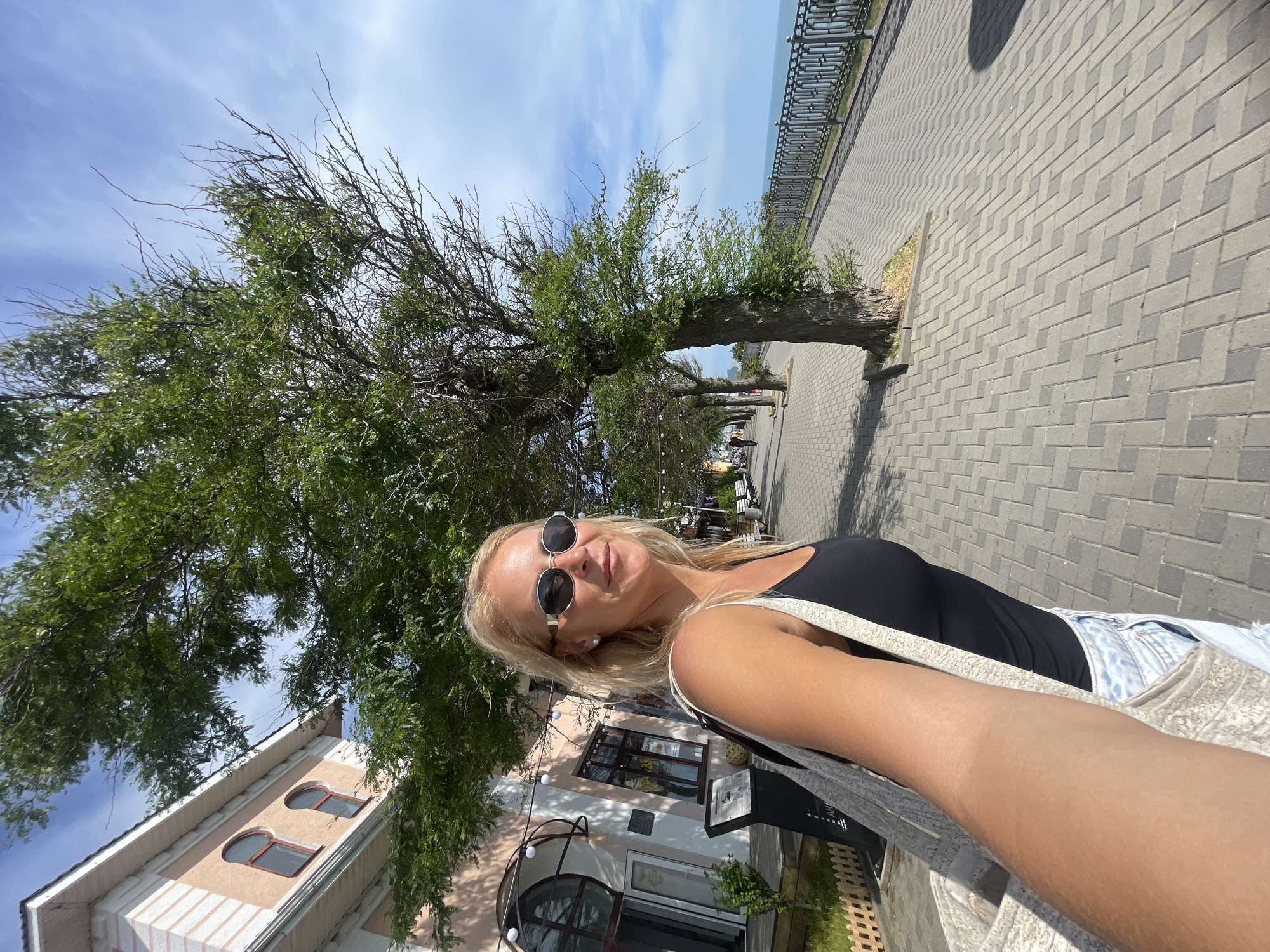 A blonde woman with sunglasses, wearing a black tank top and denim shorts, takes a selfie on a paved promenade in Anapa, Russia. She is carrying a light-colored tote bag over her shoulder. The promenade is lined with trees and benches, and to her right, a metal railing separates the walkway from the sea. In the background, a building with arched windows and decorative trim is visible. The sky is blue with some clouds, and the sun is shining, casting shadows.
