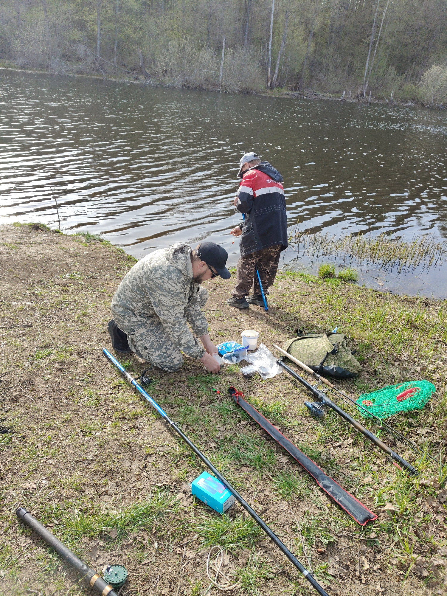 Two people are fishing by a lake. One person is kneeling on the ground, preparing fishing gear. The other person is standing by the water's edge, holding a fishing rod. There are several fishing rods and other fishing equipment lying on the ground. The lake is surrounded by trees and grass. The location is in a natural area, presumably outside of a city.