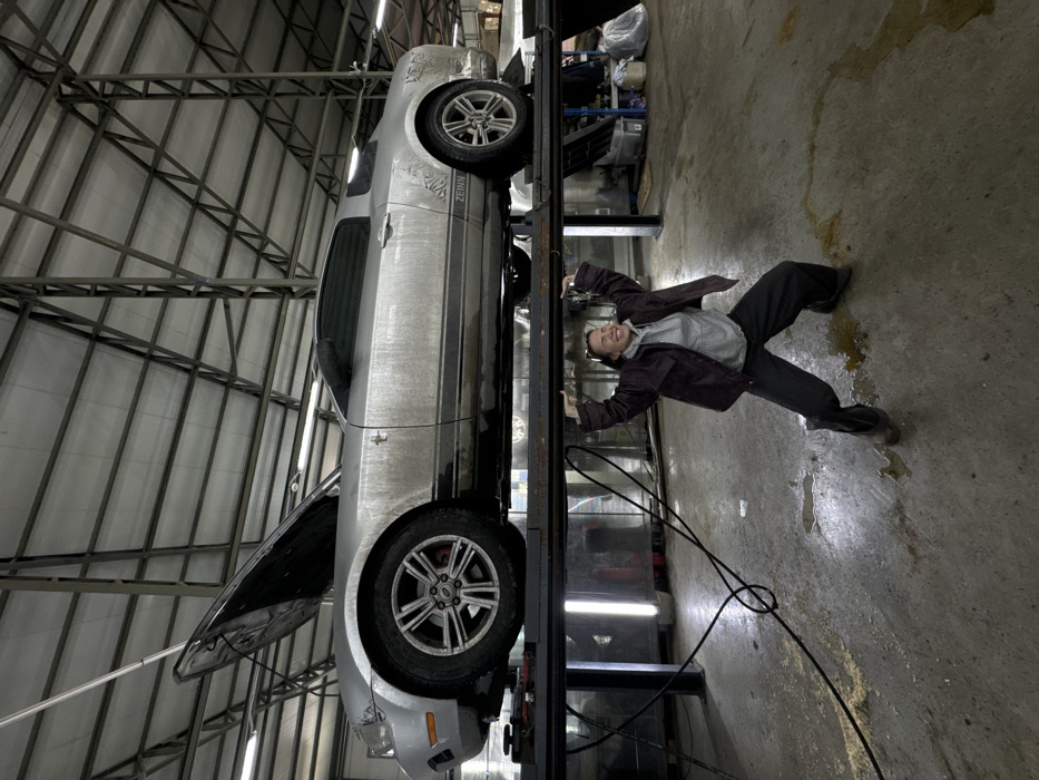 The scene depicts an interior view of a vehicle service bay located in Zhabkino, Russia. A silver Ford Mustang, visibly soiled with grime, is elevated on a dark-colored hydraulic car lift, positioned centrally within the frame. The vehicle's hood is open. Directly beneath the elevated lift, a person with dark hair, dressed in a grey hoodie, a dark jacket, dark trousers, and boots, stands with arms raised and hands pressed against the underside of the lift's platform, adopting a pose suggesting physical support of the vehicle and lift. The individual displays a wide, smiling facial expression. The concrete floor exhibits multiple wet patches and streaks, with a thick black cable coiled nearby. The facility features a high ceiling constructed with exposed metal trusses and illuminated by industrial fluorescent lights. Various workshop equipment and storage units are visible in the background.