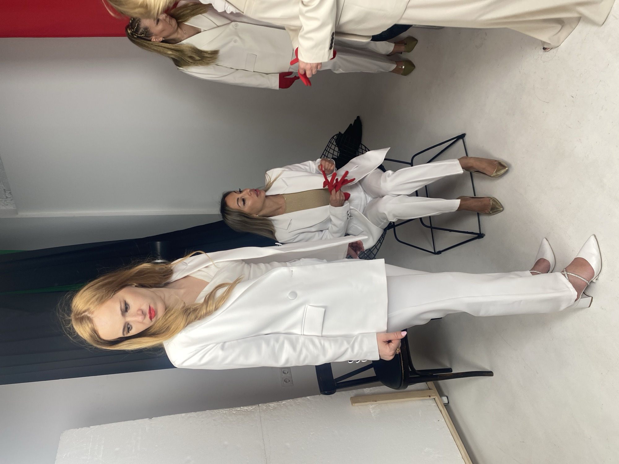 The image depicts an indoor studio setting, possibly in Sochi, Russia, with a white wall and floor. Four women are present, all dressed in white suits. In the foreground, a woman with blonde hair looks down and to her left. To her right, another woman is seated on a chair, holding a pair of red gloves. Further back, two other women are visible, also wearing white suits and one of them is holding red gloves. The scene suggests a photo shoot or a fashion-related event.
