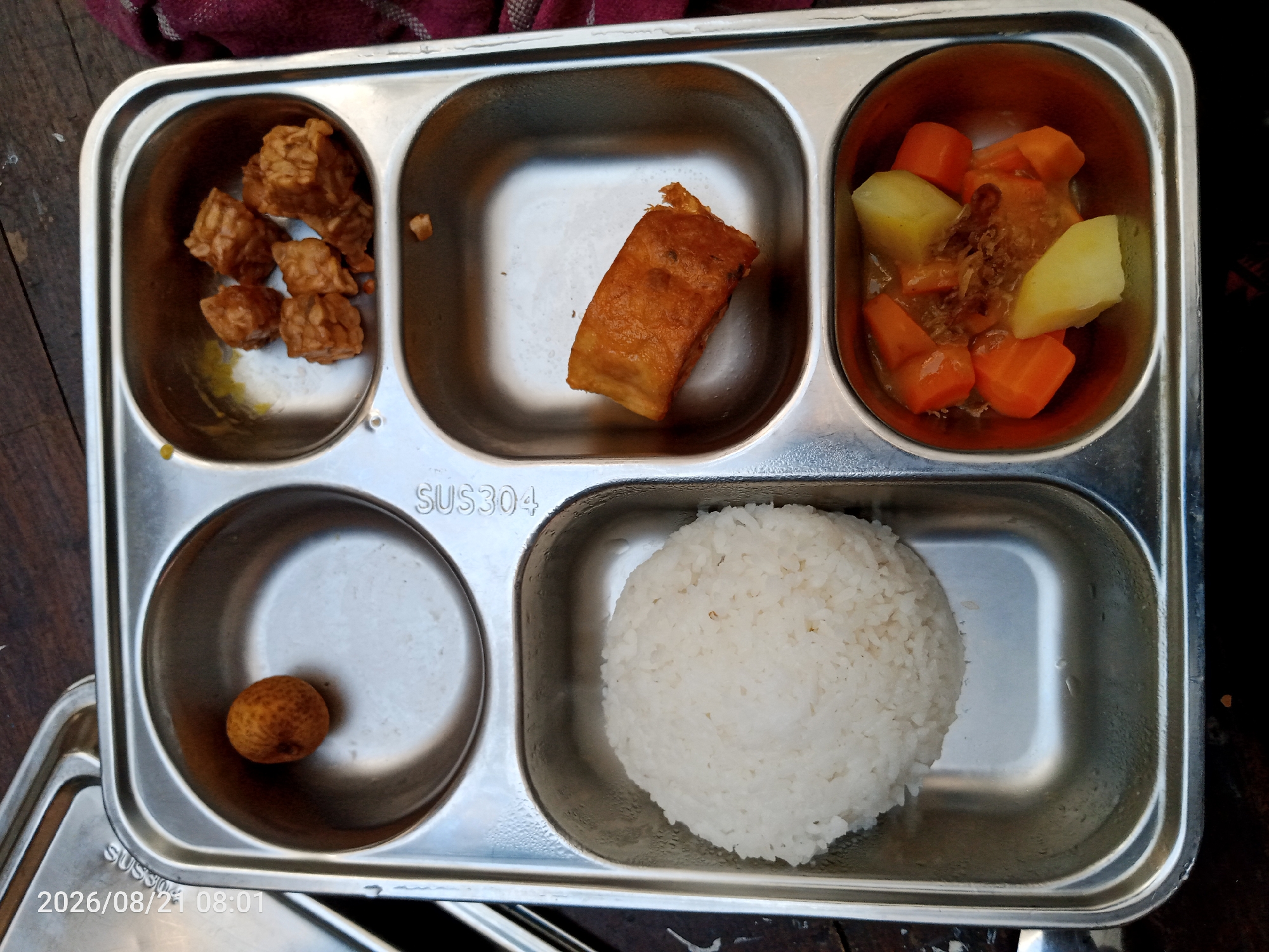A metal compartment tray, labeled "SUS304", holds a meal. The tray is placed on a wooden surface, and a dark red fabric is visible in the upper left corner. The meal consists of several portions. In the top left compartment are several pieces of fried tempeh. Adjacent to this, in the top center compartment, is a single piece of fried food, possibly a piece of tofu or fried dough. The top right compartment contains a stew with diced carrots and potatoes, topped with fried shallots. The bottom left compartment holds a single, round, brown fruit, possibly a longan. The bottom right compartment contains a mound of white rice. A timestamp "2026/08/21 08:01" is visible in the lower left corner of the image. This meal is likely from a canteen or food stall in Surakarta, Indonesia, given the commonality of these food items and the labeling on the tray.