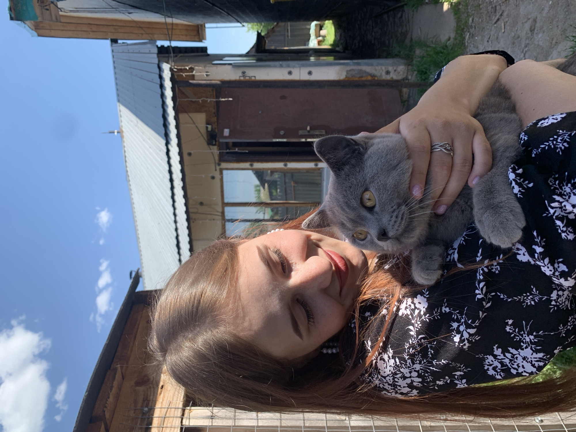 A woman is holding a grey cat. The woman has long brown hair, fair skin, and is wearing a black and white patterned dress. She is smiling with her eyes closed. Her left hand is resting on the cat. The cat has yellow eyes. The background is a mix of buildings, including a corrugated metal structure, wooden structures, and a narrow passage or alley. The sky is blue with some clouds visible. The location is Фоки, Russia.