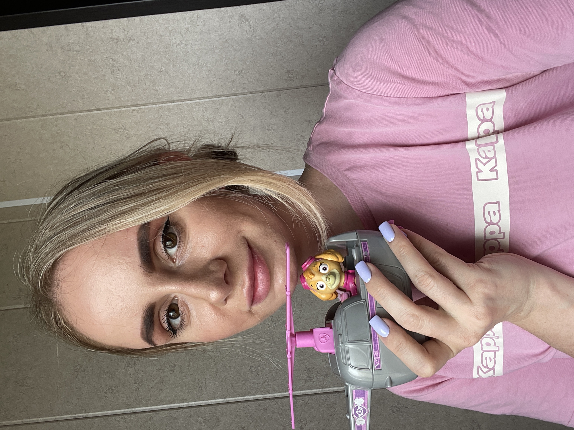 A fair-skinned woman with blonde hair holds a Paw Patrol Skye toy helicopter. She wears a pink shirt with white Kappa lettering. The background is a tiled wall. There is no indication of Vladivostok, Russia.