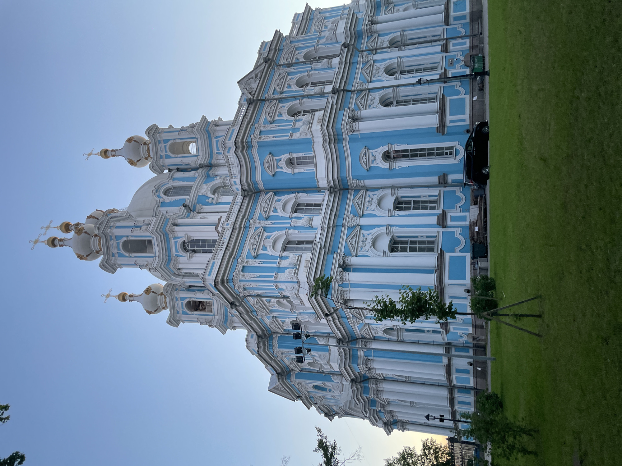 The image shows the Smolny Cathedral, a prominent landmark in Saint Petersburg, Russia. The cathedral is a magnificent example of Baroque architecture, characterized by its striking blue and white facade, ornate detailing, and gilded onion domes. The building is bathed in soft, natural light, suggesting it was taken during the early morning or late evening.
In the foreground, a lush green lawn stretches out, with a few young trees and bushes adding to the serene atmosphere. A black minivan is parked on the paved area in front of the cathedral, and a lamppost stands nearby. The sky above is a clear, pale blue, indicating fair weather conditions.
The Smolny Cathedral is a part of the Smolny Institute, which was originally founded as a women's educational institution. The cathedral itself is an active place of worship and a popular tourist destination.
