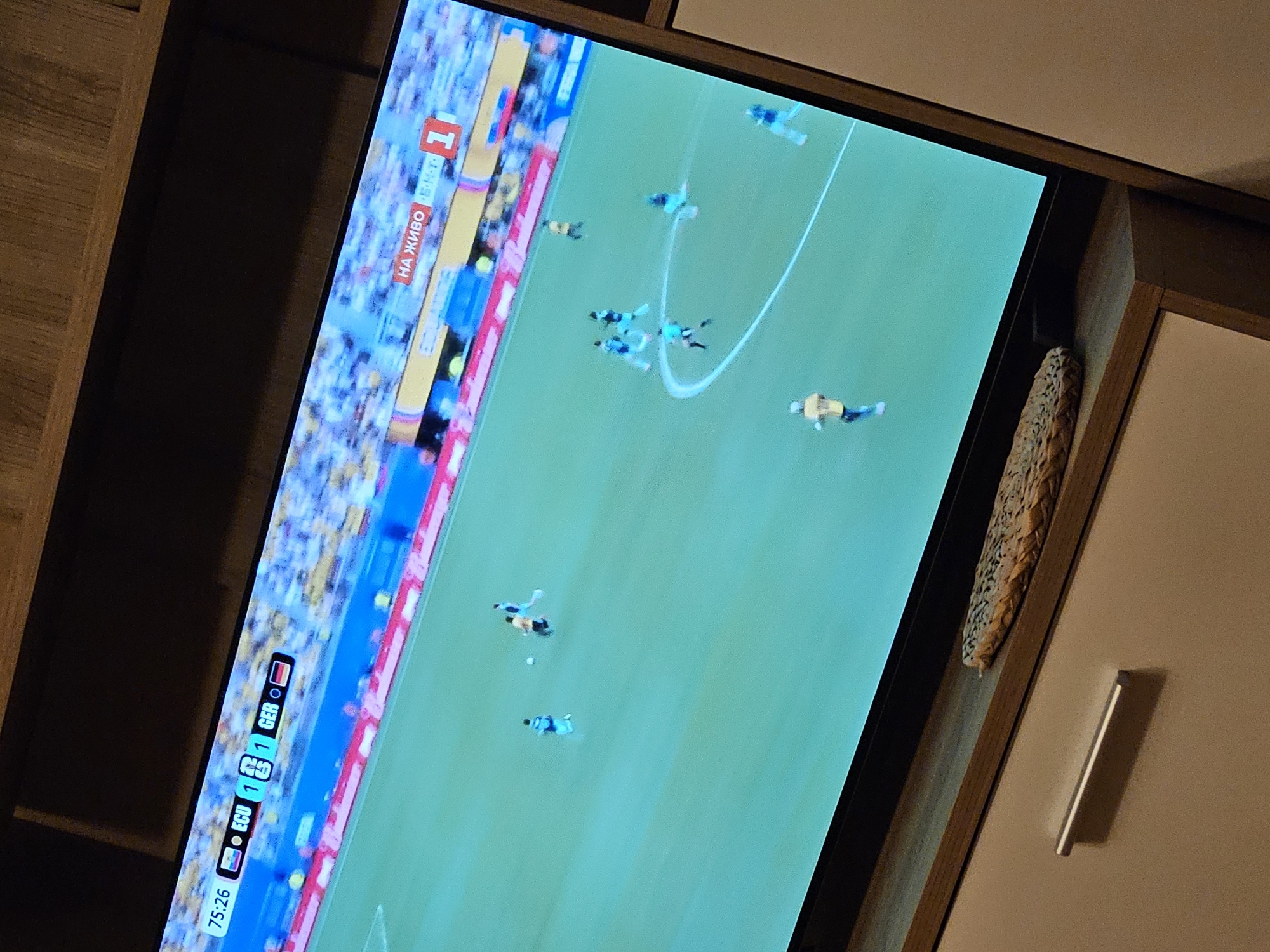 The image shows a television screen displaying a soccer match. The scene is a vibrant green soccer field with several players in blue and yellow uniforms scattered across it. A white soccer ball is visible near the center of the field. The broadcast includes a score overlay at the bottom left, indicating the time is 75:26 and the score is 1-1 between ECU and GER. To the left of the score, a banner reads "НА ЖИВО 1" in red and white text, suggesting the match is live. The stadium appears to be filled with spectators, though they are blurred in the background. The overall setting is an indoor environment, with part of a cabinet visible to the right of the television screen. The lighting suggests it is likely daytime, but the indoor setting makes it difficult to determine the exact time of day. No specific emotions are discernible from the blurred figures of the players.