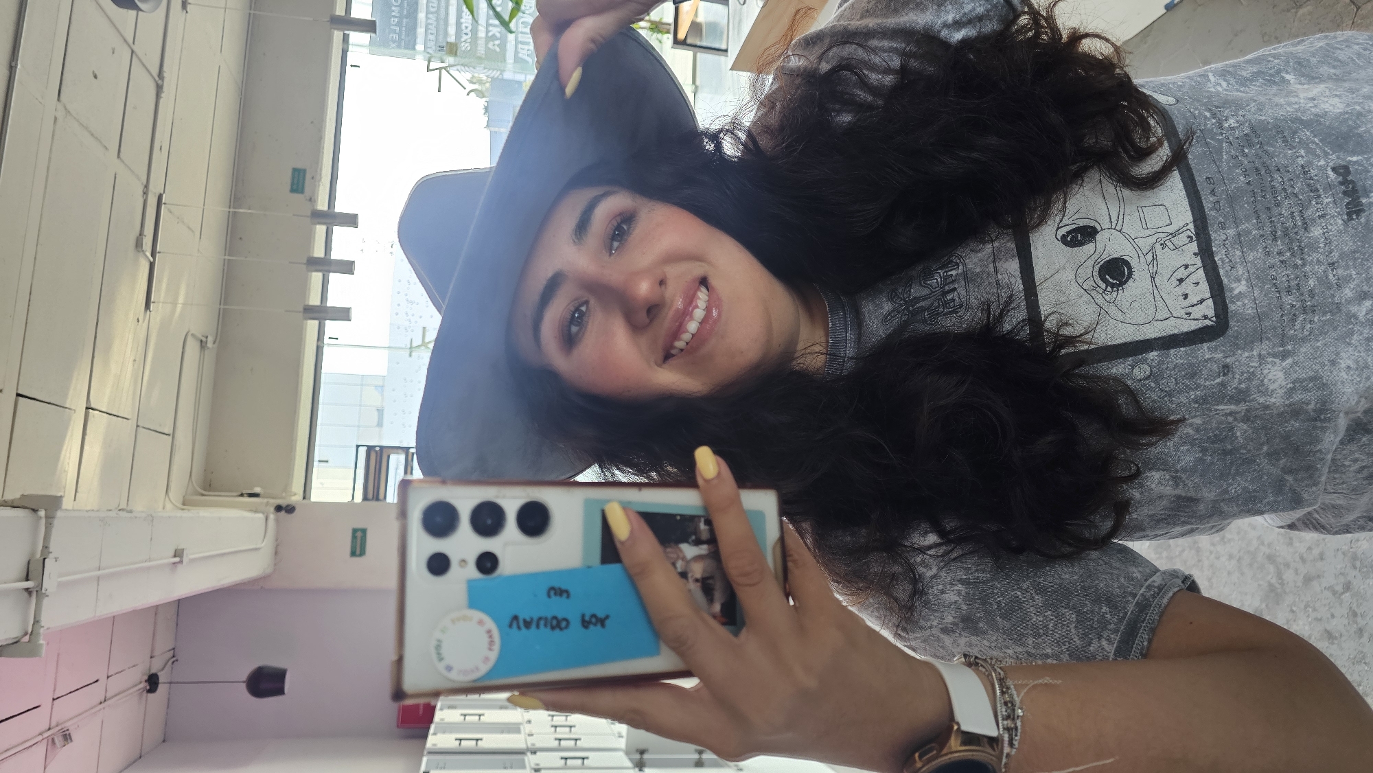 A woman with long dark hair, wearing a gray hat and a gray t-shirt with a graphic design, is holding a smartphone with a blue sticky note and a photograph stuck to the back of it. She is smiling and appears to be taking a selfie. She is wearing a watch. The background shows a window with bright daylight and what seems to be an interior space with white walls and some lighting fixtures. Based on the sticker visible on the phone case, it is likely that the photo was taken in Puebla, Mexico.