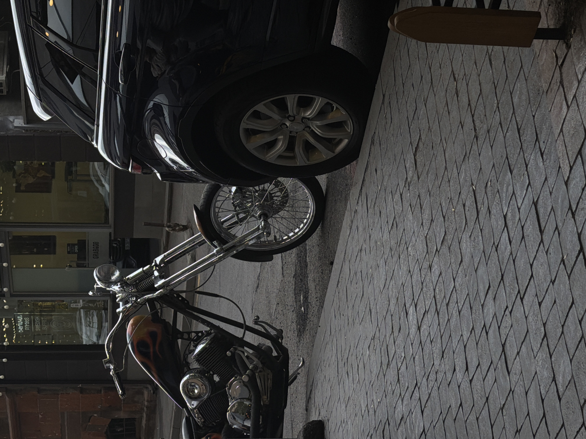 Captured at an oblique angle, the image reveals a section of a street scene. A black automobile is partially visible at the top, adjacent to a segment of a brick walkway that runs along the sidewalk. A custom-designed motorcycle stands out, featuring a vibrant, flame-painted fuel tank. The backdrop includes the exterior of a building with expansive glass storefronts. The signage "GRAB&GO" is noticeable, as is a window display promoting a "SWEDISH BUFFET." The scene appears to be a street in Yerevan, Armenia, as indicated by the local business names and architecture.