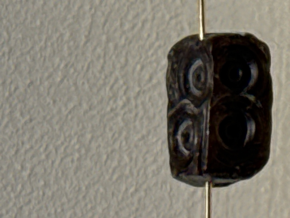 The scene depicts a single, dark, roughly rectangular bead with rounded edges, centered against a light-colored, textured background. The bead, possibly made of stone or glass, measures approximately twice as wide as it is tall and exhibits intricate carved or molded spiral patterns on its visible surfaces; specifically, two prominent double-spiral motifs are present on its upper face. A thin, golden-colored metallic wire pierces the bead horizontally, extending from both its left and right sides. No individuals are present. The environment is indistinct beyond the textured background, suggesting an indoor display or studio setting. This object is located in Kamariotissa, Greece.